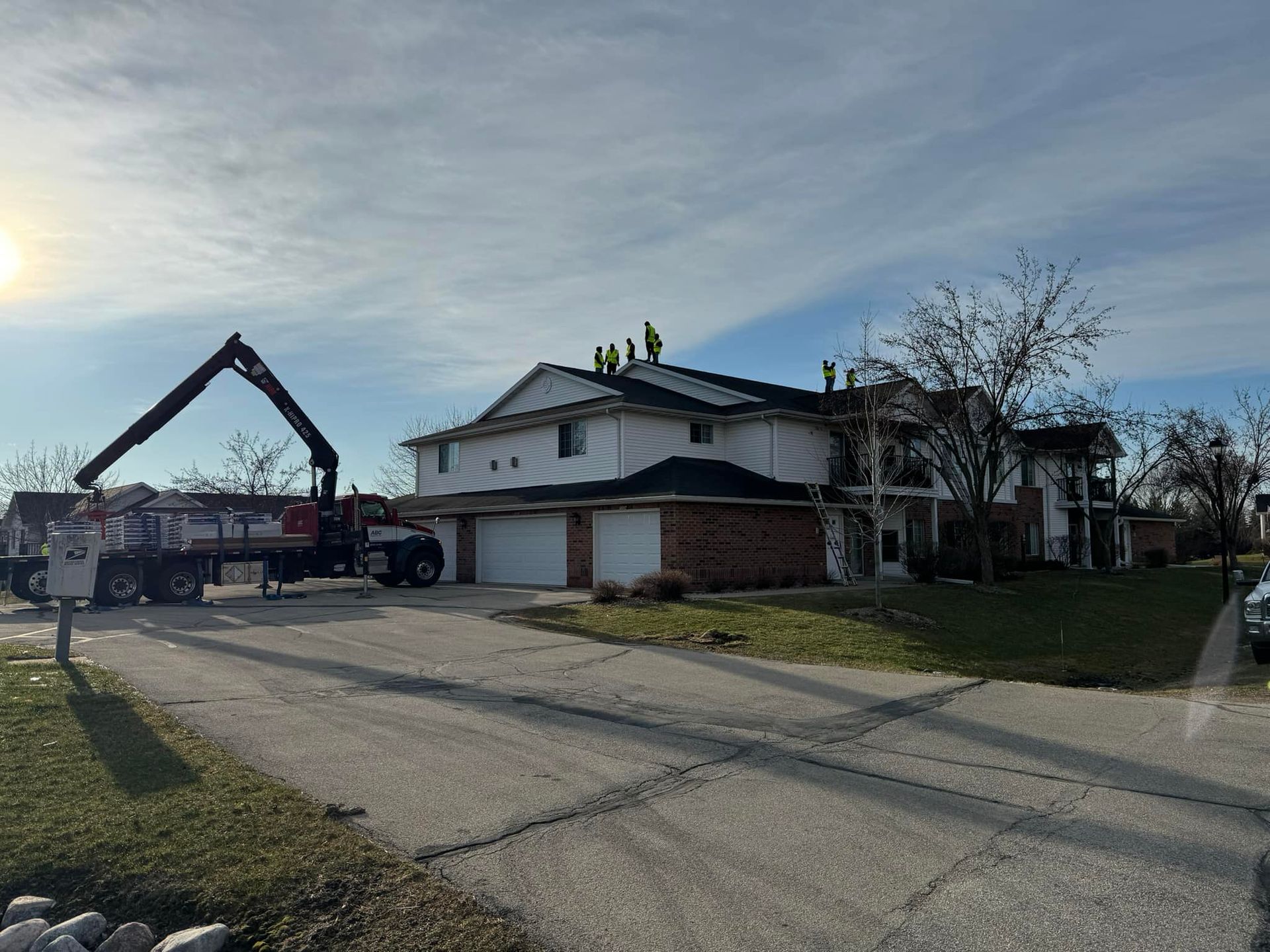 A crane truck parks in front of a white two-story suburban house while construction workers perform roof repairs.