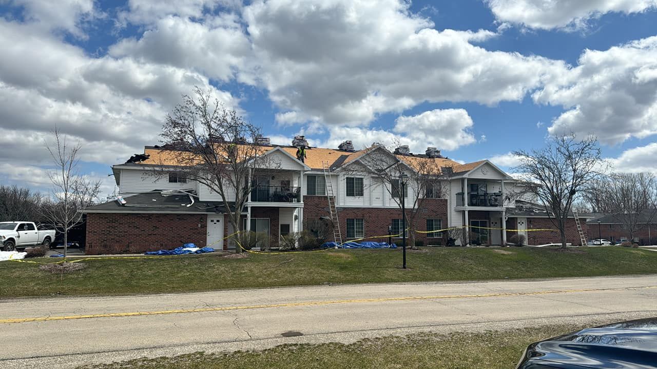 An apartment building with a partially damaged, exposed roof sits under a partly cloudy blue sky.