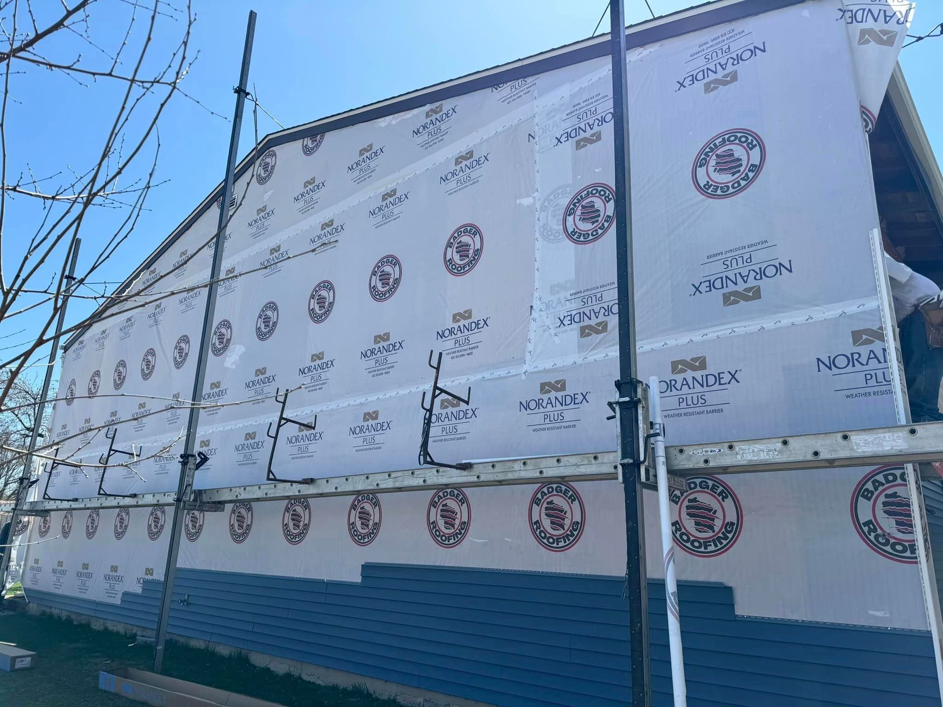 A house exterior under construction with blue siding on the lower half and protective building wrap on the upper walls.