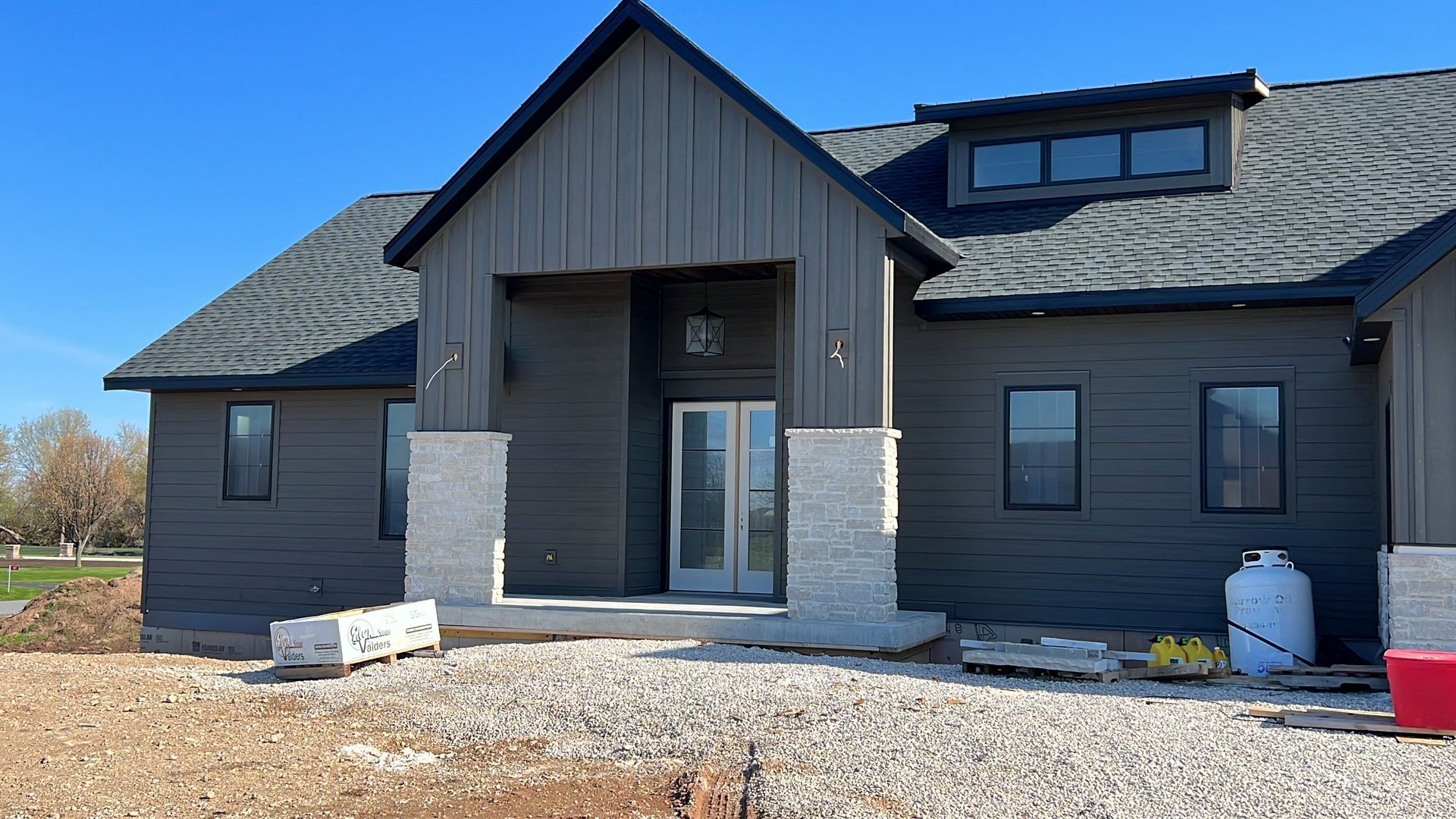 Modern dark-gray home exterior under construction with stone pillar accents, a gravel driveway, and a clear blue sky.