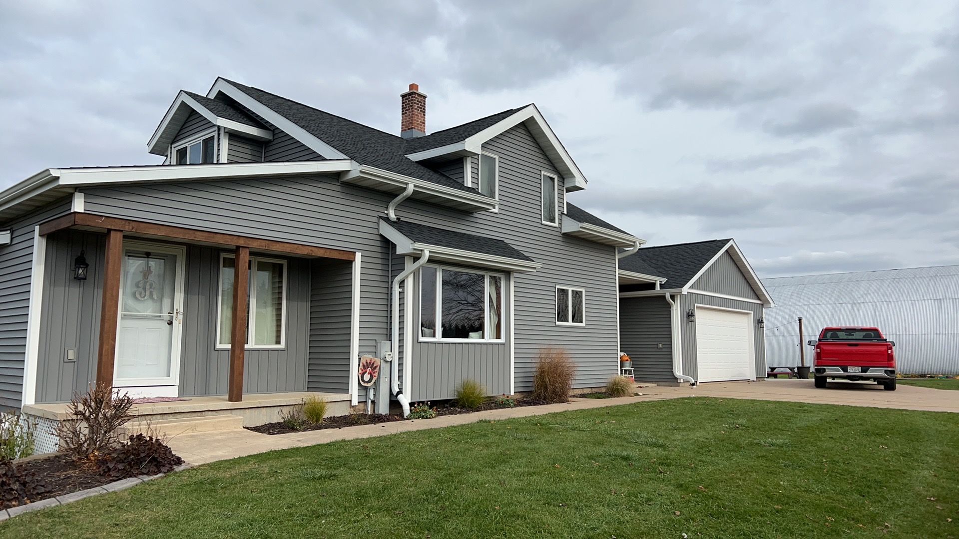 A two-story grey farmhouse with a dark roof and attached garage, situated on a grassy lawn with a red truck nearby.