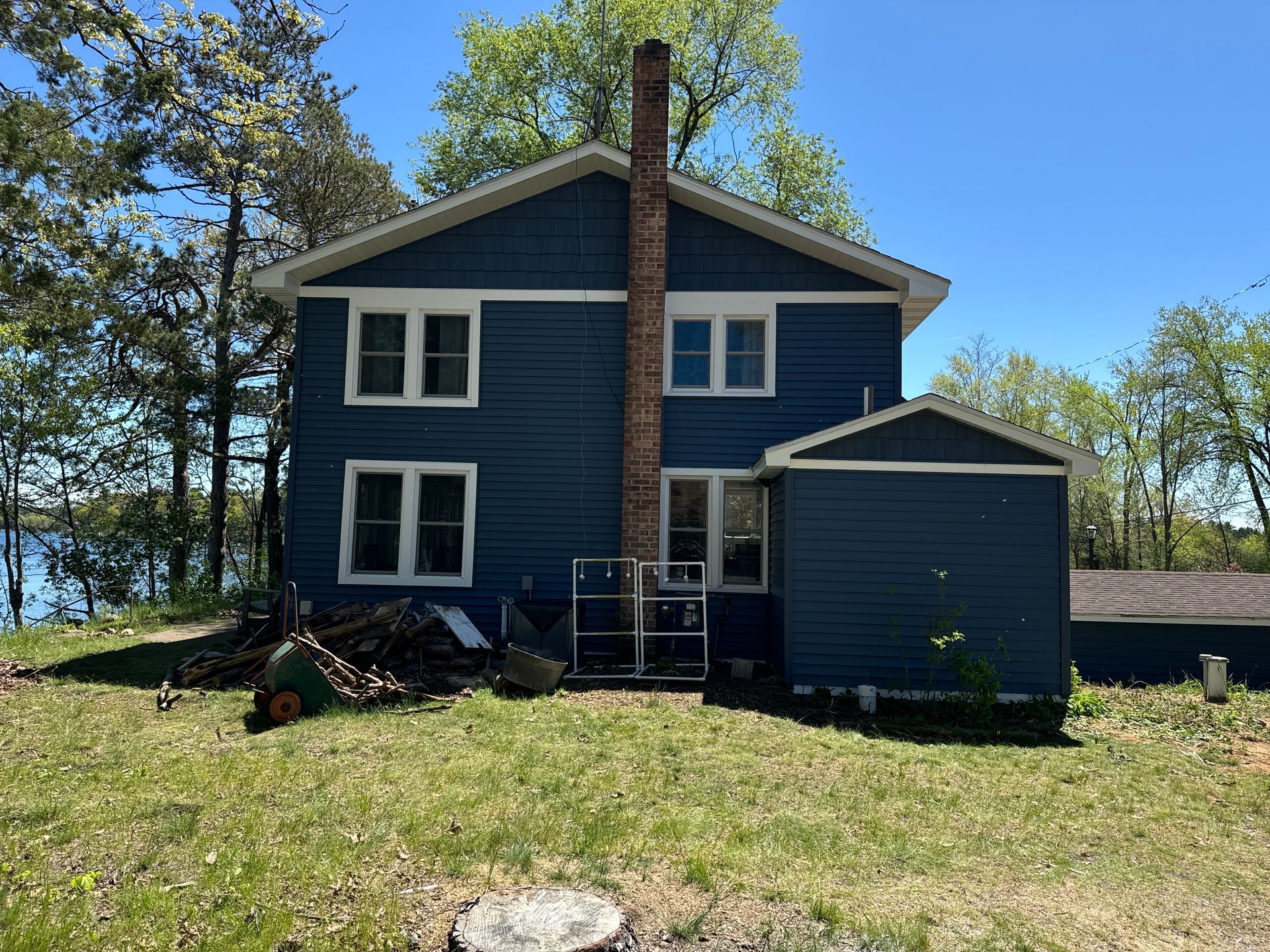 A two-story blue house with a brick chimney, situated on a grassy lot with trees and a lake in the background.