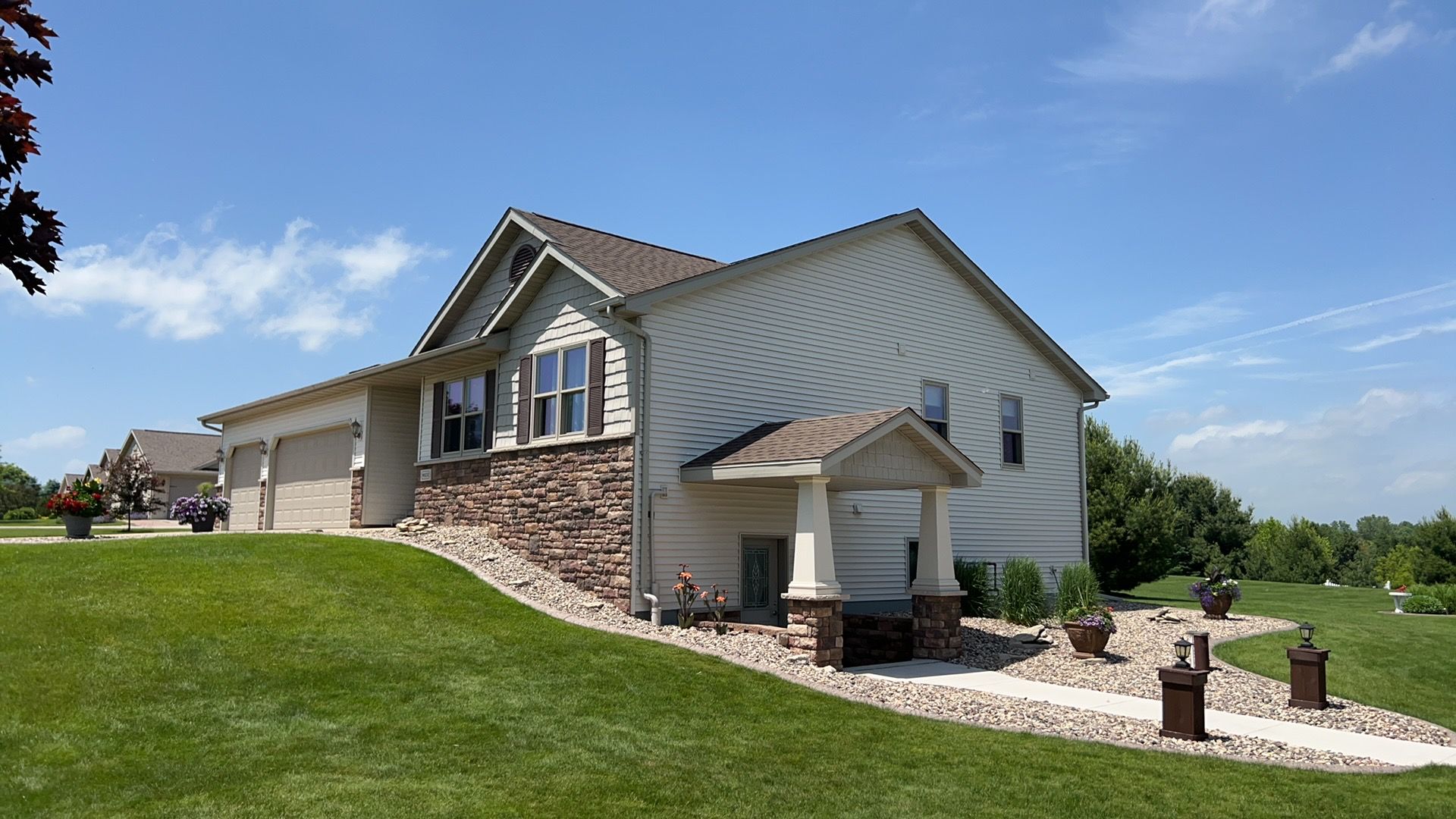 A single-story suburban house with beige siding, a stone facade, and a covered entryway set on a grassy hill.