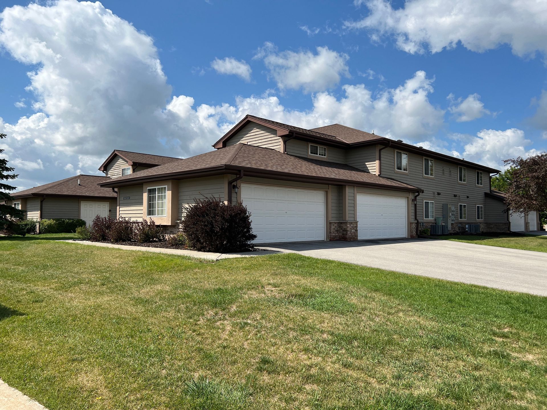 A two-story, tan-colored townhouse complex with two white garage doors, surrounded by a grassy lawn under a blue sky.