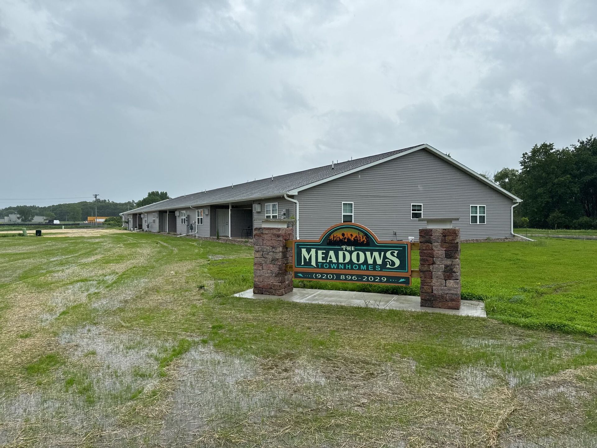 A sign for The Meadows at Stonefield in front of a long, gray building on a grassy lot under a cloudy sky.