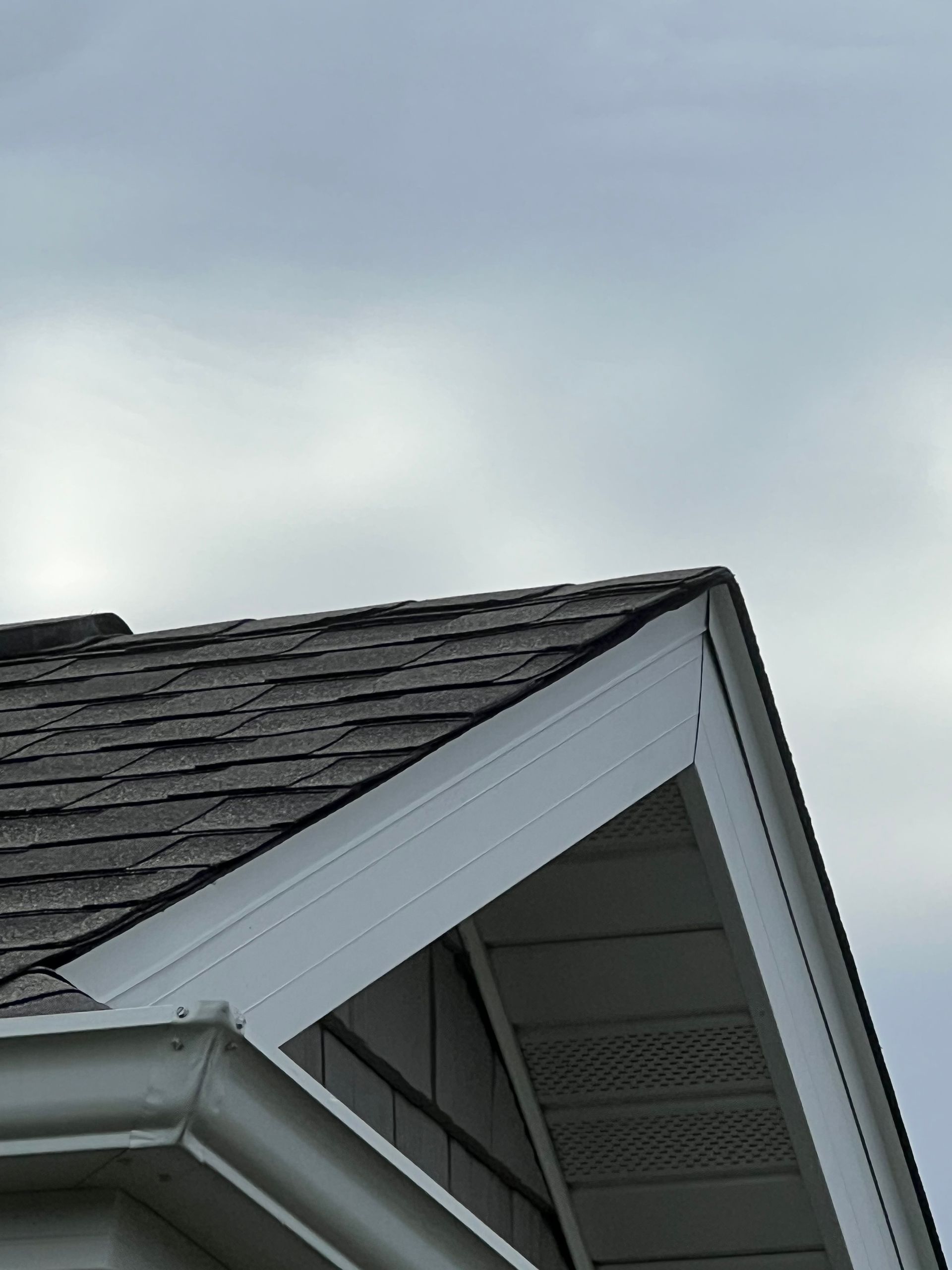 A low-angle view of a grey shingled roof gable with white trim against an overcast sky.