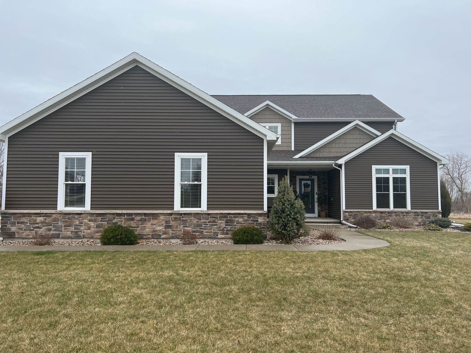 A two-story house with dark gray horizontal siding and a stone veneer base under a cloudy sky with a front yard.