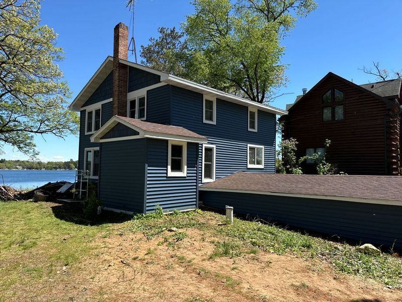 A two-story blue house with a brick chimney sits on a grassy shore next to a lake, with a dark wood house nearby.