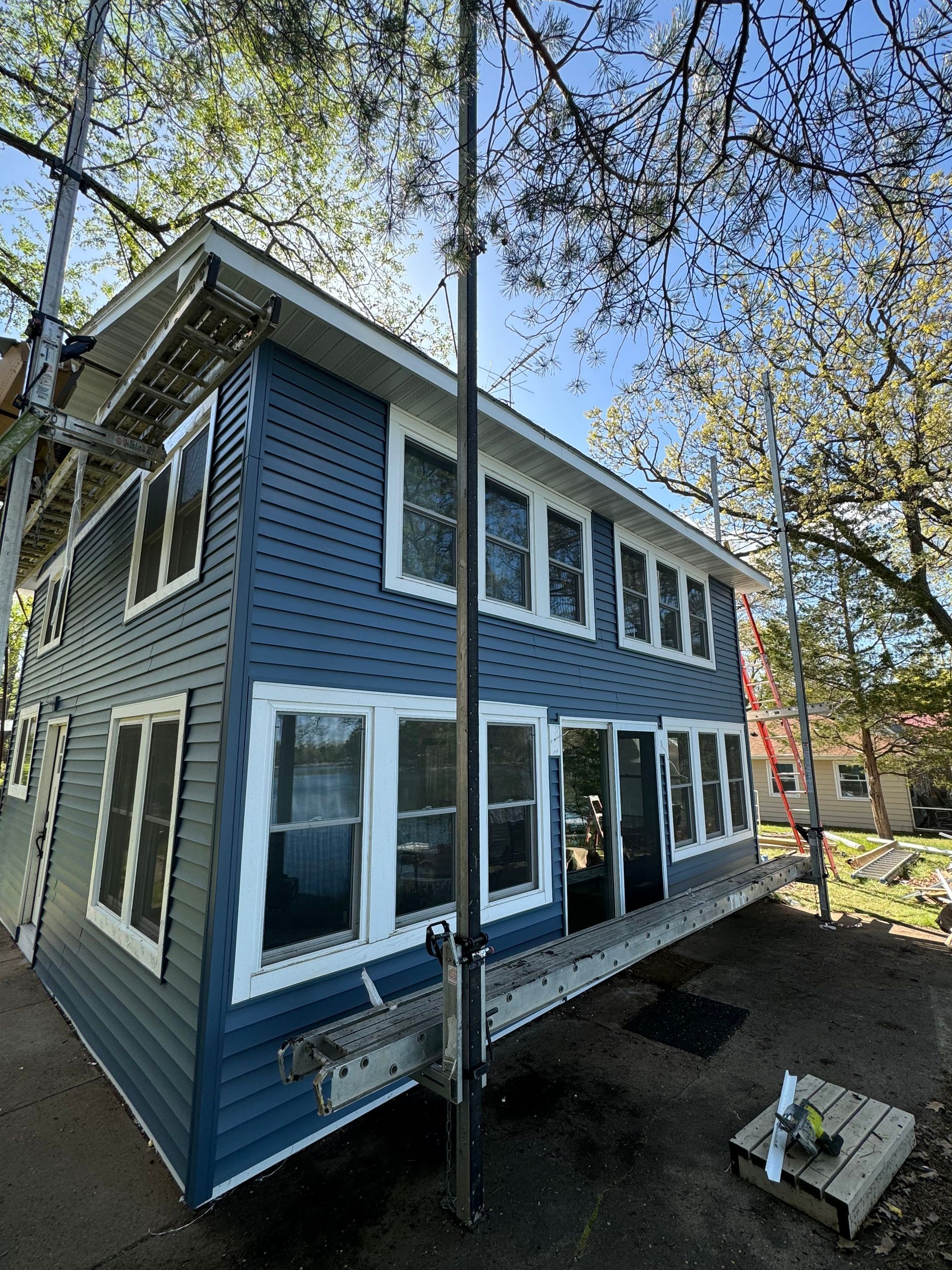 A two-story blue house under renovation with scaffolding around the exterior and bare trees under a clear blue sky.