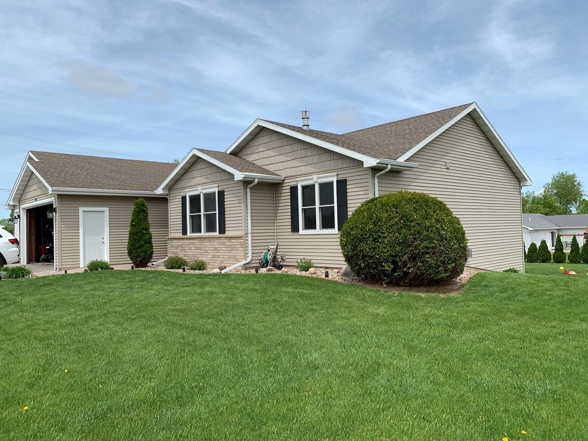 A one-story suburban home with beige siding, a brown shingled roof, a front lawn, and a large round shrub.