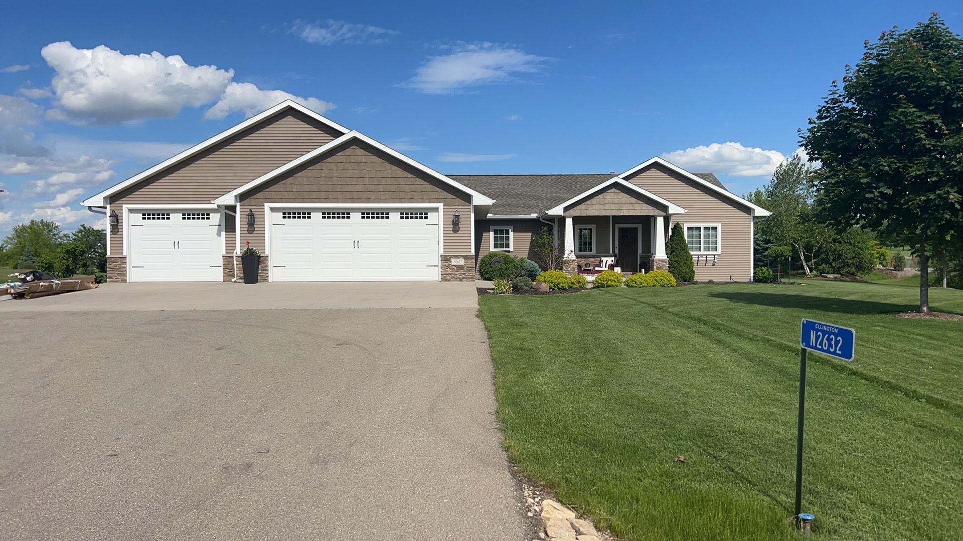 A tan, single-story house with a three-car garage and a wide driveway under a blue, cloudy sky.