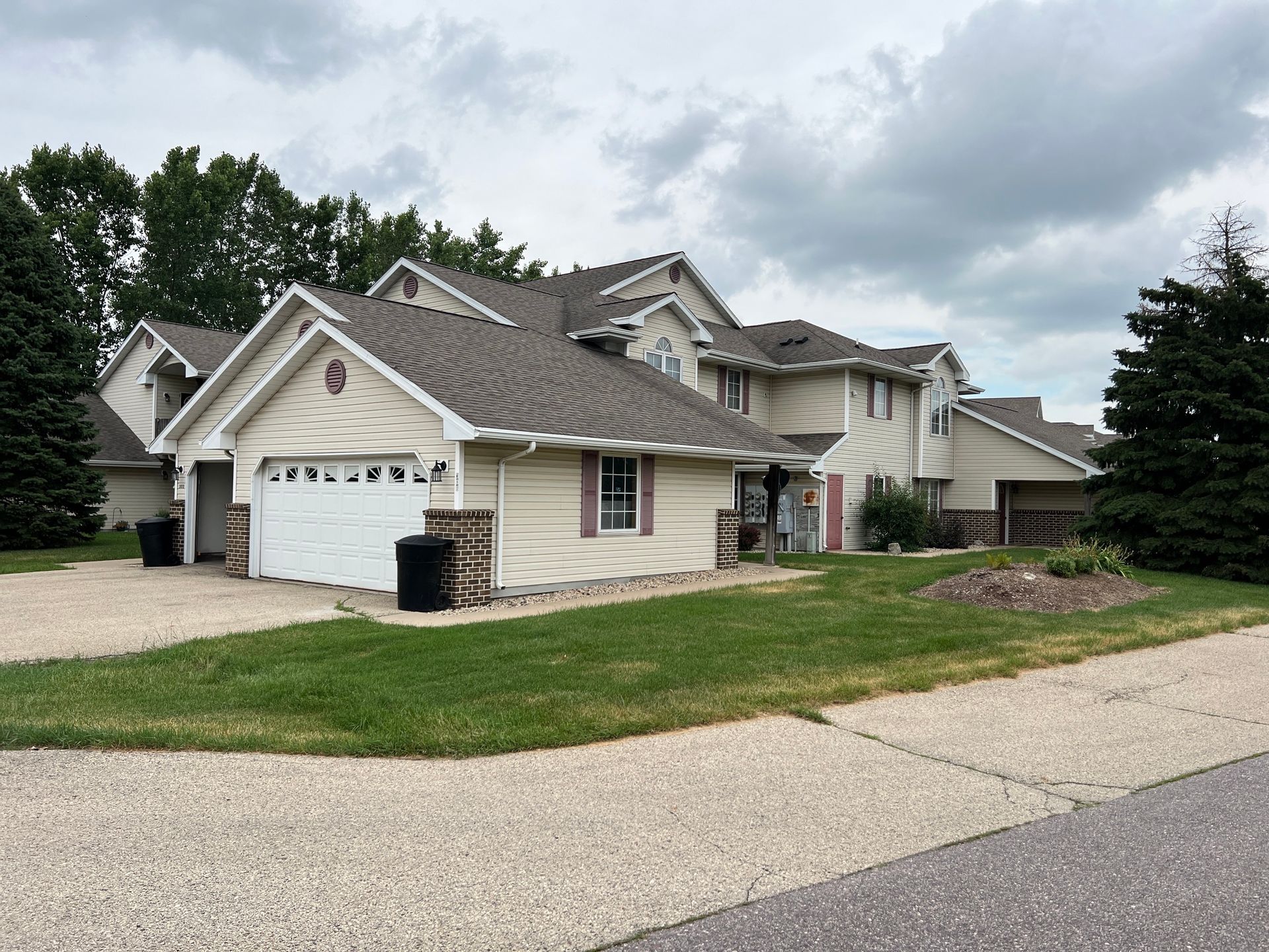 A tan, multi-story suburban house with a white garage door, surrounded by green lawns and pine trees under a cloudy sky.