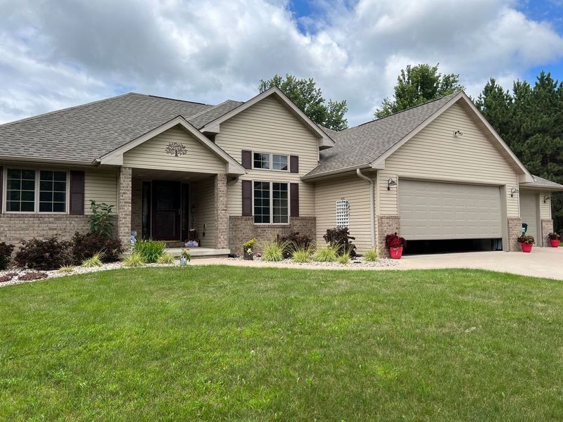 A tan house with brick accents, a gray shingled roof, a covered entryway, and a multi-car garage on a lawn.