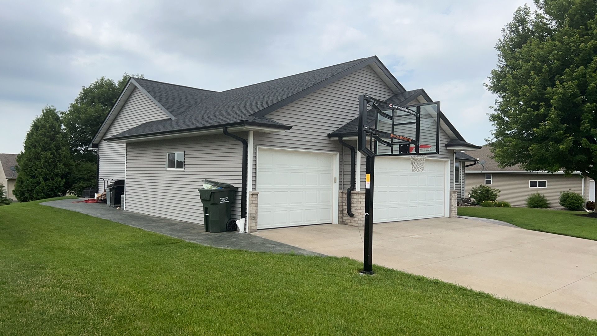 A house with beige horizontal siding, a black roof, a two-car garage, and a basketball hoop on the concrete driveway.