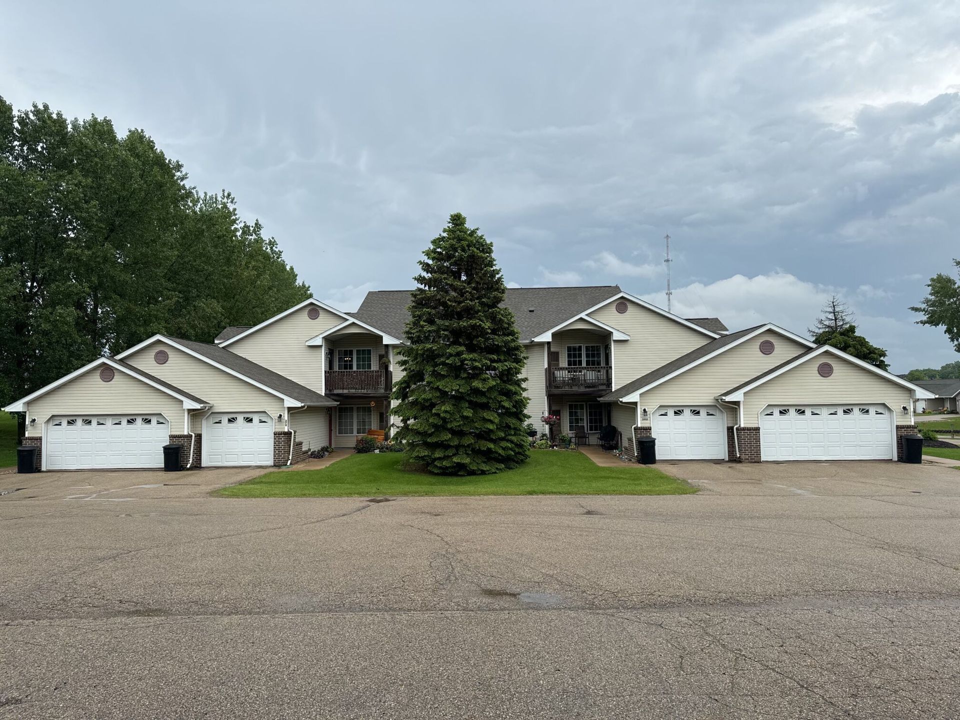 A two-story tan apartment building with four white garage doors and a large evergreen tree in the front lawn.