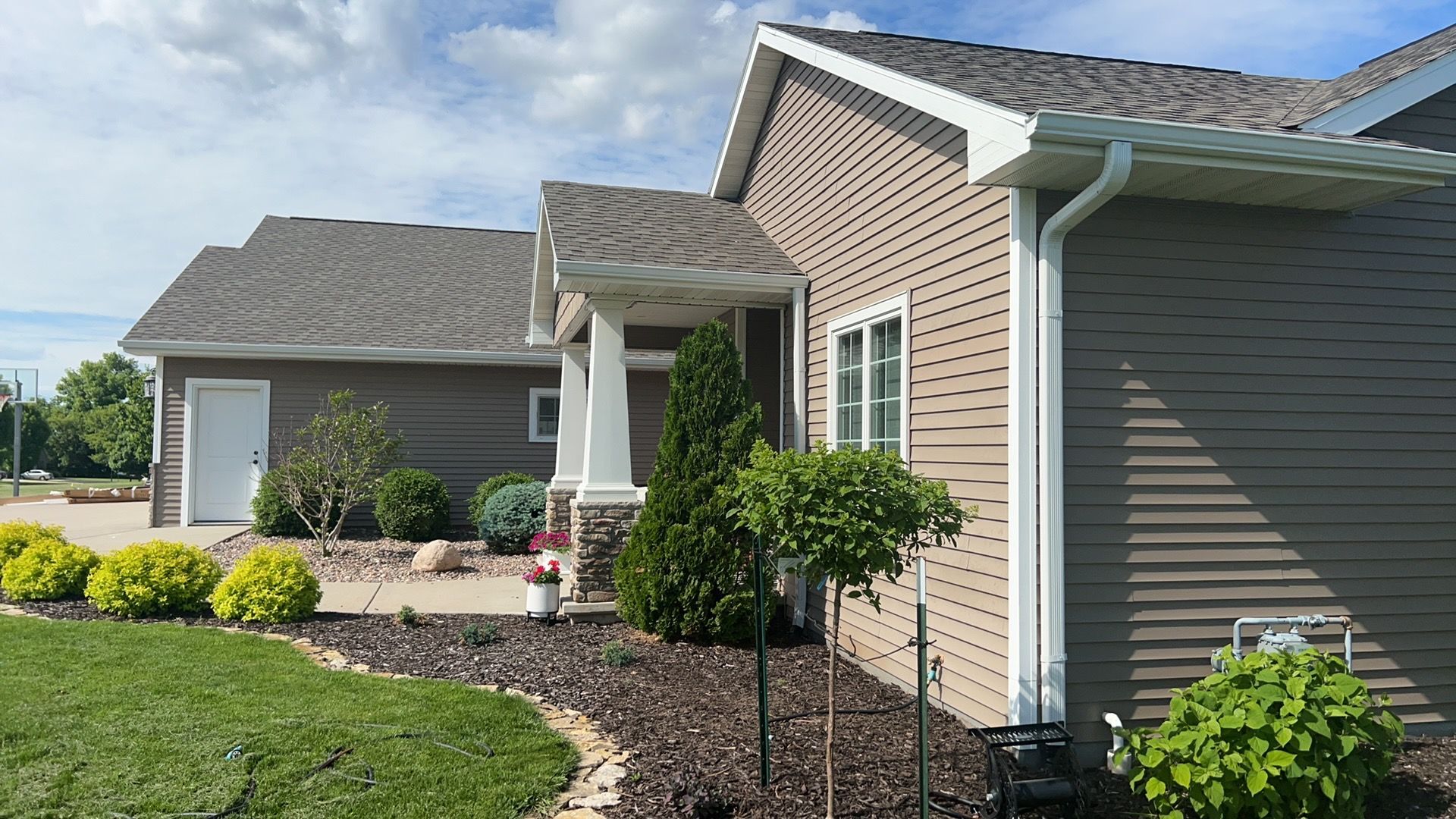 A beige house with a covered entryway and tan siding, featuring a green lawn, shrubs, and mulch beds on a sunny day.