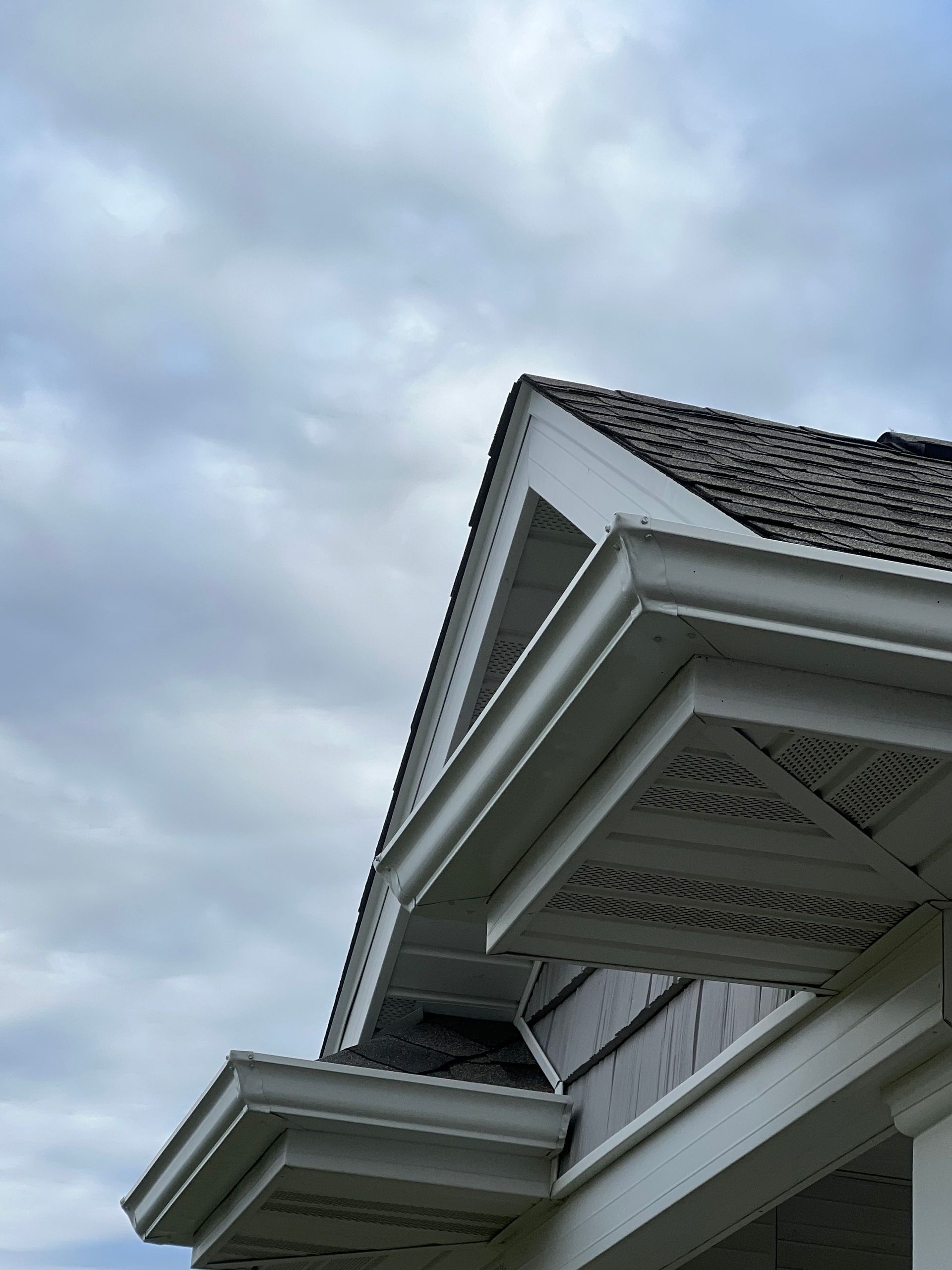 Low-angle view of the white trim, dark roof shingles, and soffits on the exterior corner of a house under a cloudy sky.
