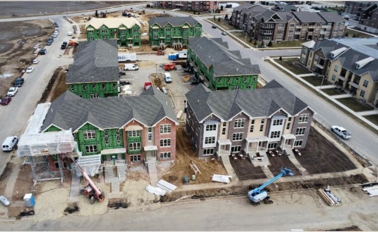 An aerial view shows several townhouses under construction, some with green structural panels, near finished units.