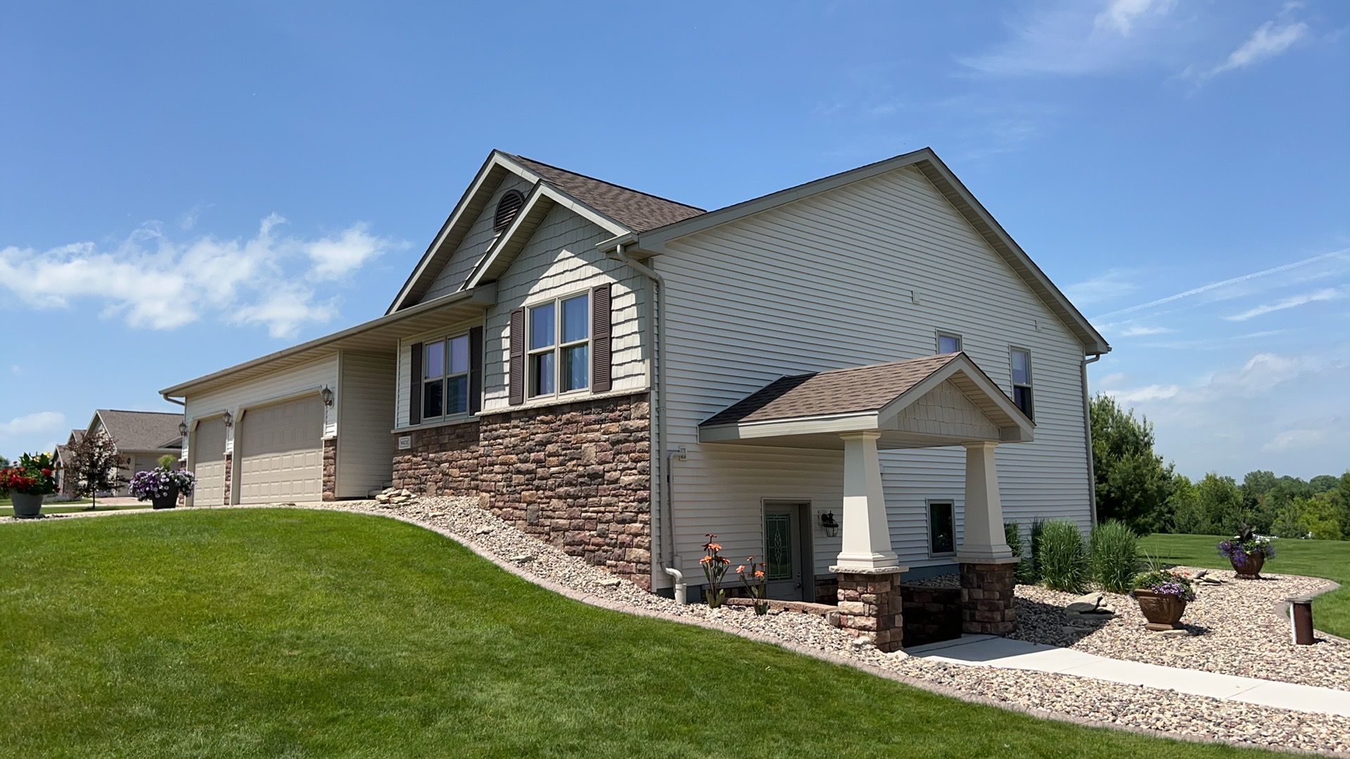 A side view of a residential house with beige siding, stone accents, and a front porch, set on a grassy lot under blue sky.