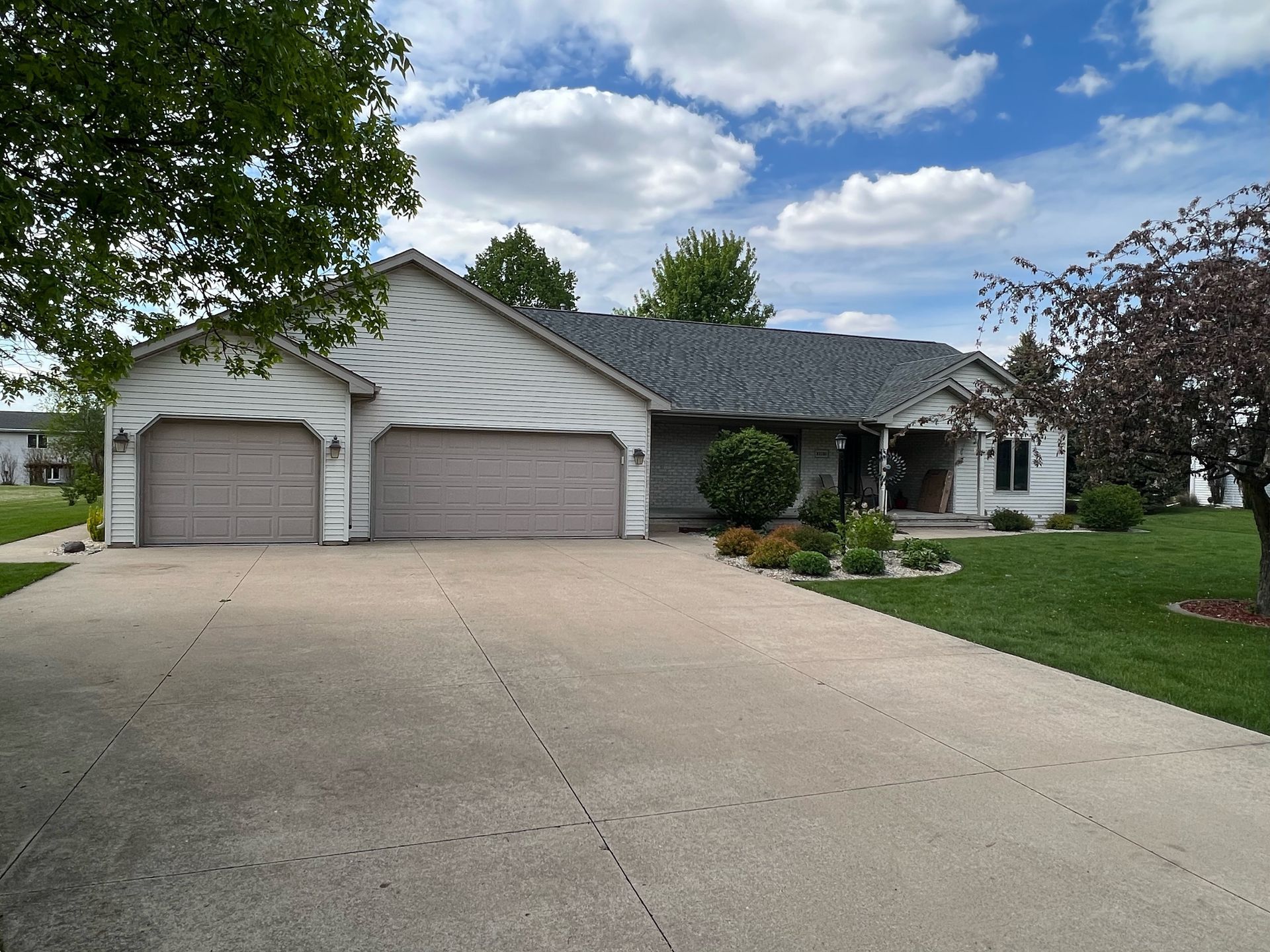 A light-colored single-story house with a three-car garage and a wide concrete driveway under a blue, cloudy sky.