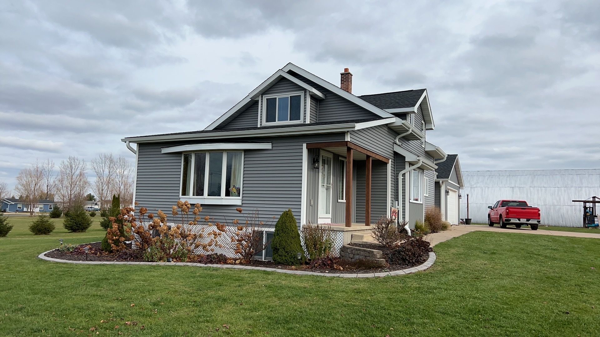 A grey, two-story house with a wooden porch and a landscaped front garden sits under a cloudy sky near a red pickup truck.