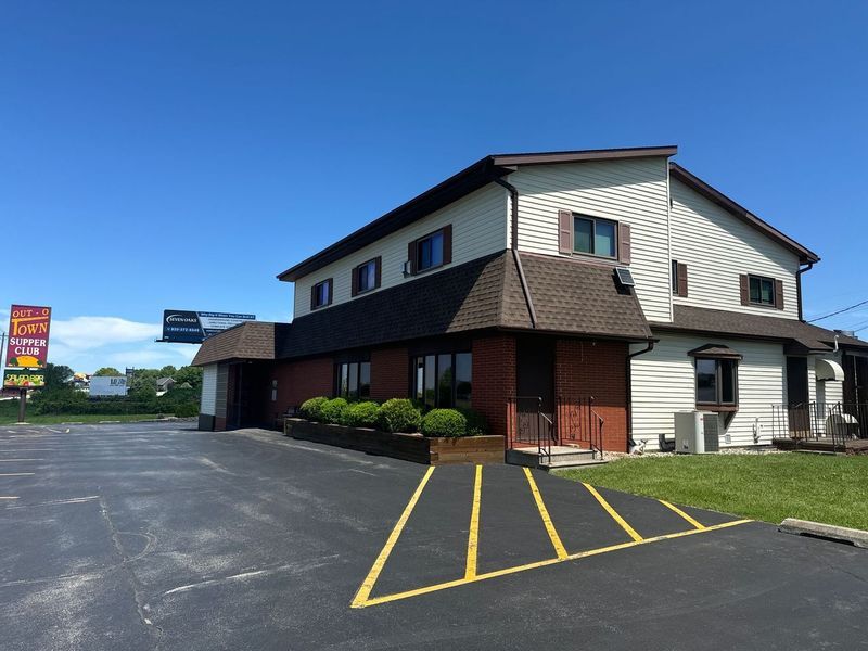 A two-story building with beige siding and brick trim sits beside an empty asphalt parking lot under a clear blue sky.