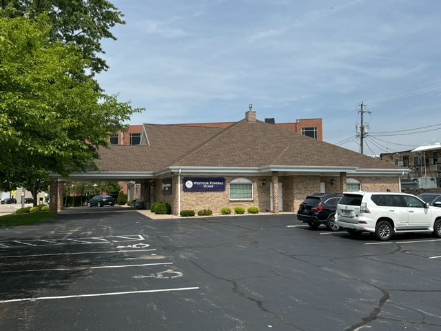 A tan brick building with a brown roof and a covered entryway, seen from a parking lot on a sunny day.