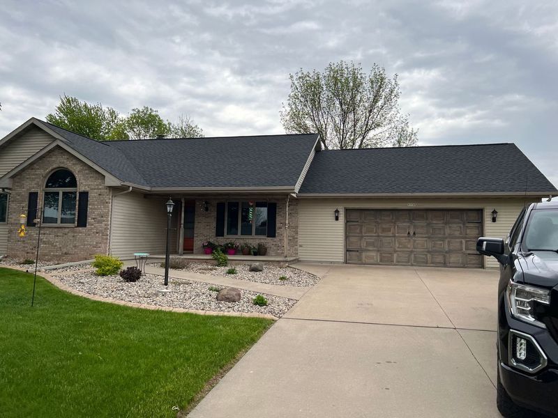 A single-story suburban house with stone and tan siding, a dark roof, a concrete driveway, and a partially visible truck.