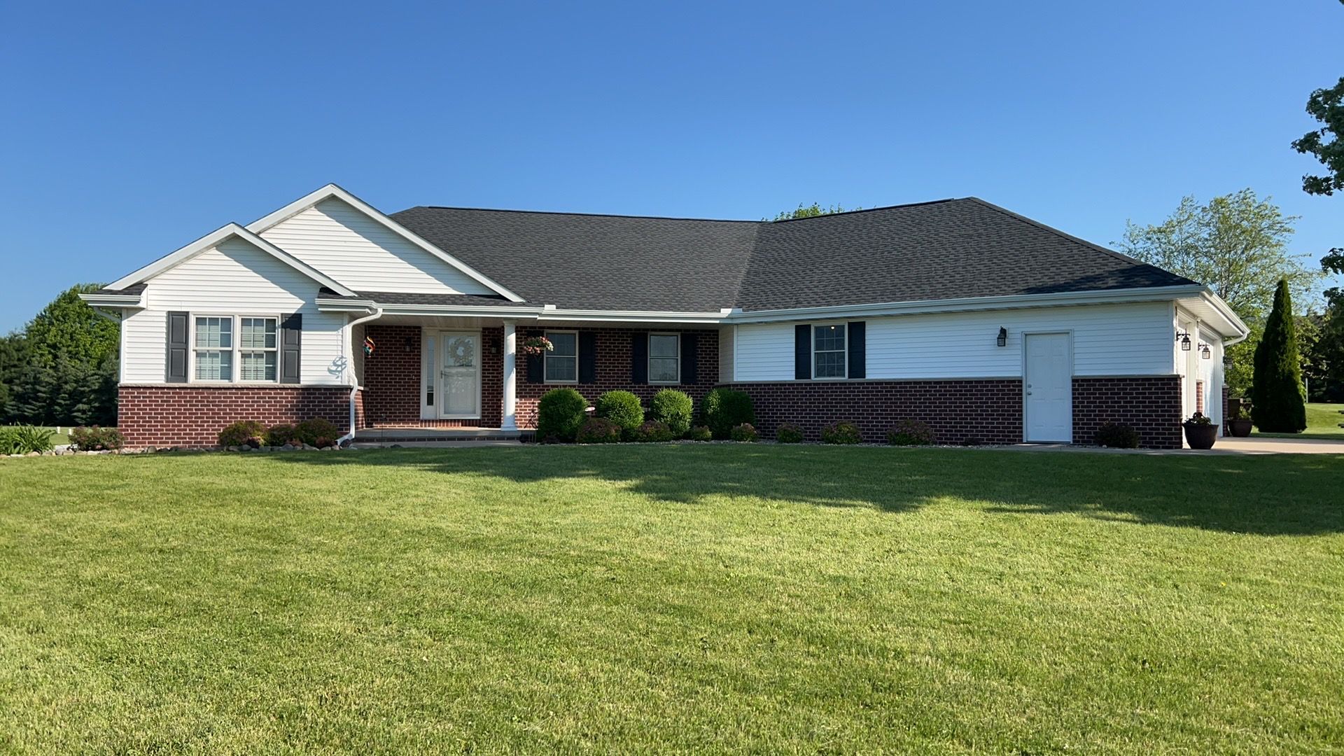A wide, one-story suburban home with a white exterior, dark red brick base, and a dark shingled roof under a clear sky.