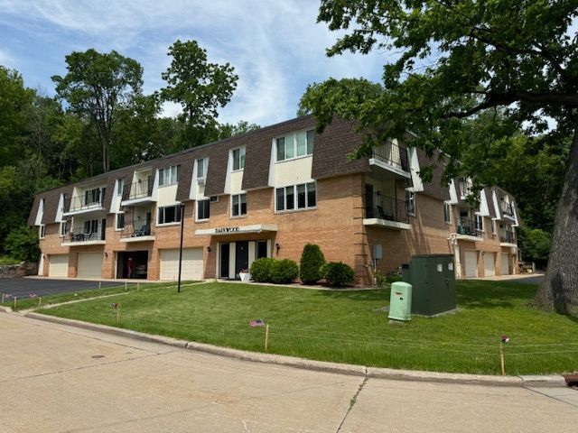 A multi-story brick apartment building with brown mansard-style roofing, balconies, and an adjacent lawn on a sunny day.