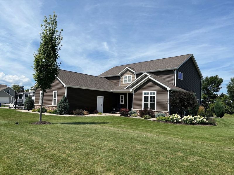 A brown two-story suburban house with a large lawn, a tall thin tree in the front yard, and a clear blue sky.