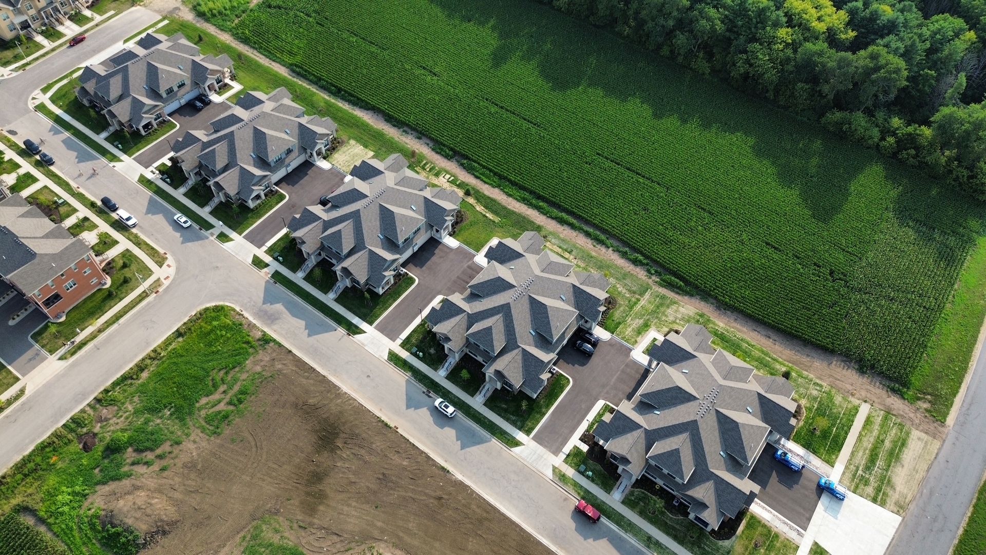 Aerial view of a suburban street lined with gray-roofed houses beside green fields and trees