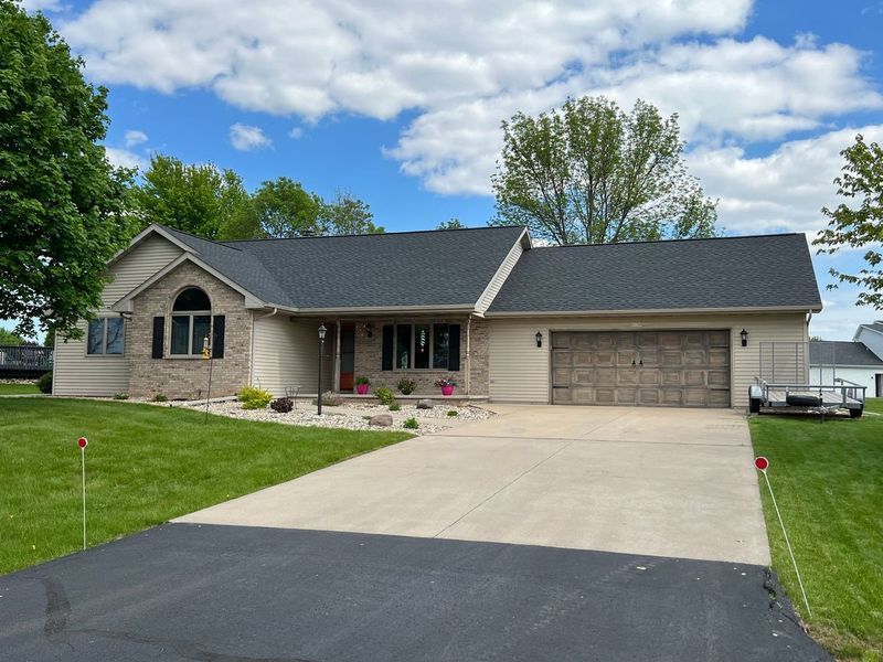 Single-story house with beige siding, stone accents, a dark roof, and a paved driveway under a blue, cloudy sky.