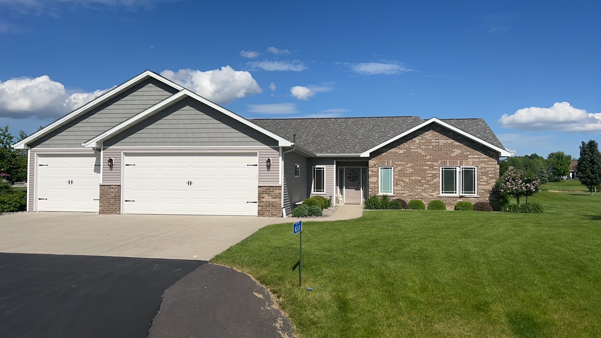 A single-story suburban house with a brick and gray siding facade, a three-car garage, and a large front lawn under blue sky.