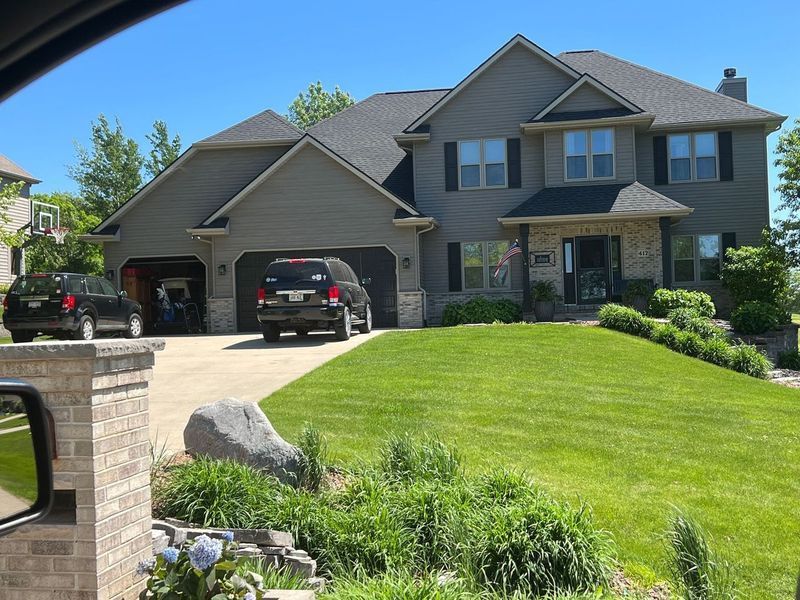 A two-story grey suburban house with a multi-car garage, manicured lawn, and two vehicles parked in the driveway.