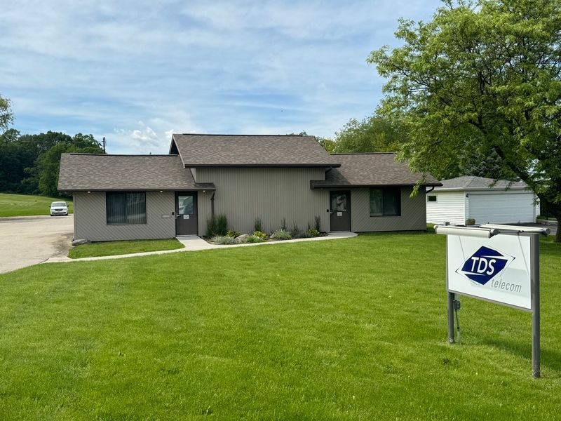 A brown, one-story commercial building with a gray roof, set on a green lawn with a white TDS sign in the foreground.