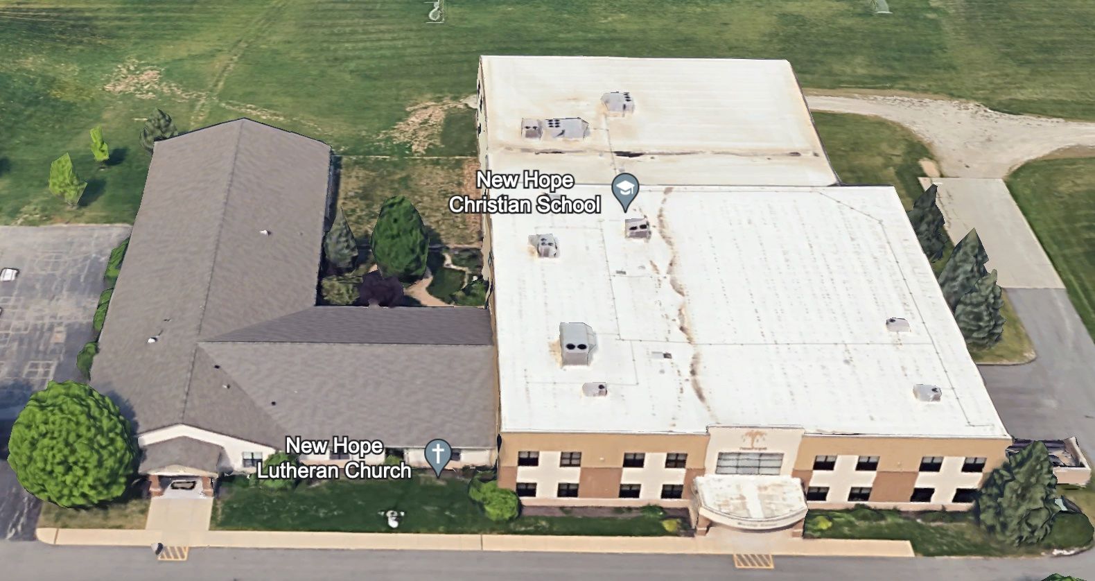 An aerial view of a school building with a brown roof section and a white flat-roofed gymnasium area on a grassy lot.