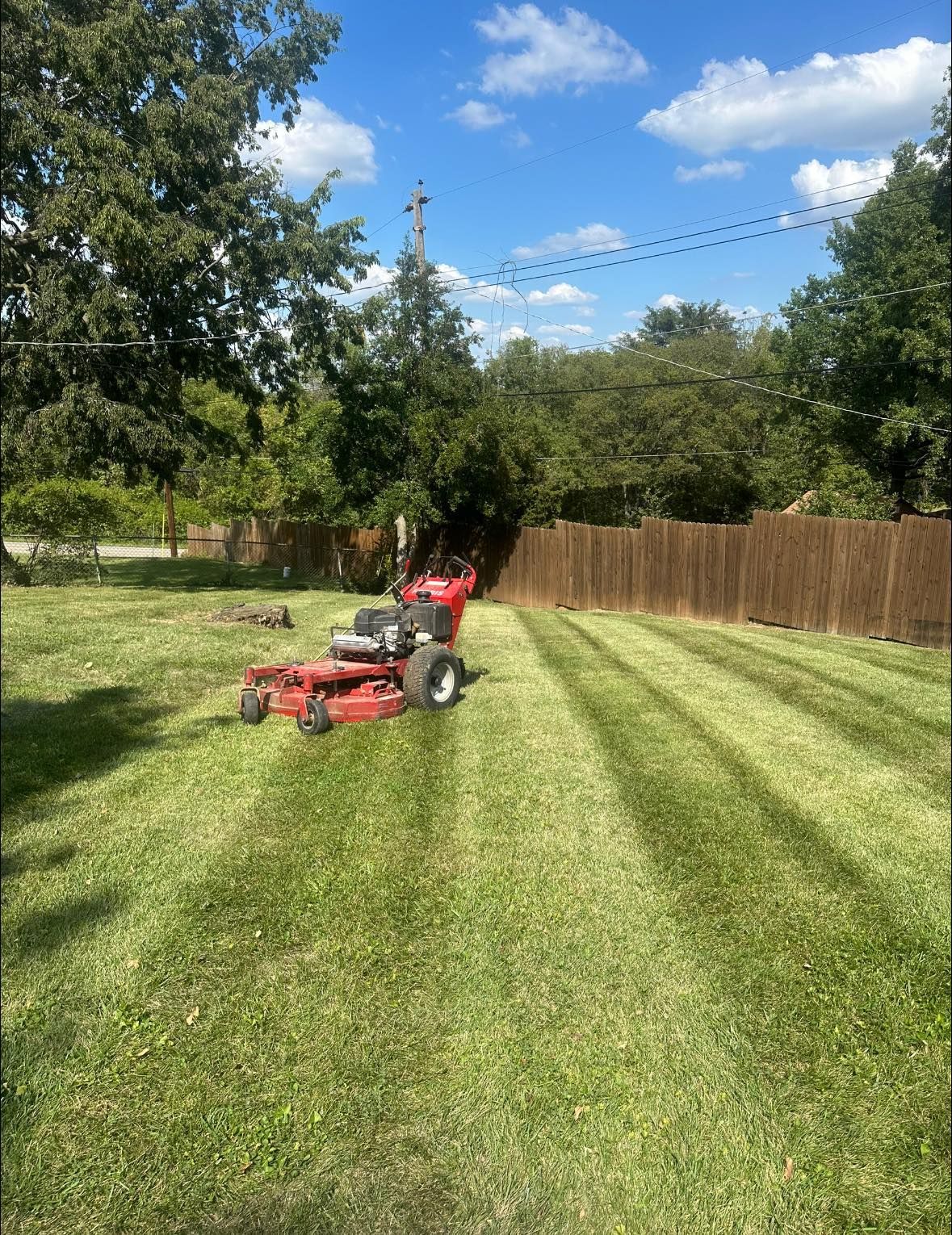 A red zero-turn mower cuts striped patterns into a green lawn in front of a wooden fence on a sunny day. A red zero-turn mower cuts striped patterns into a green lawn in front of a wooden fence on a sunny day.
