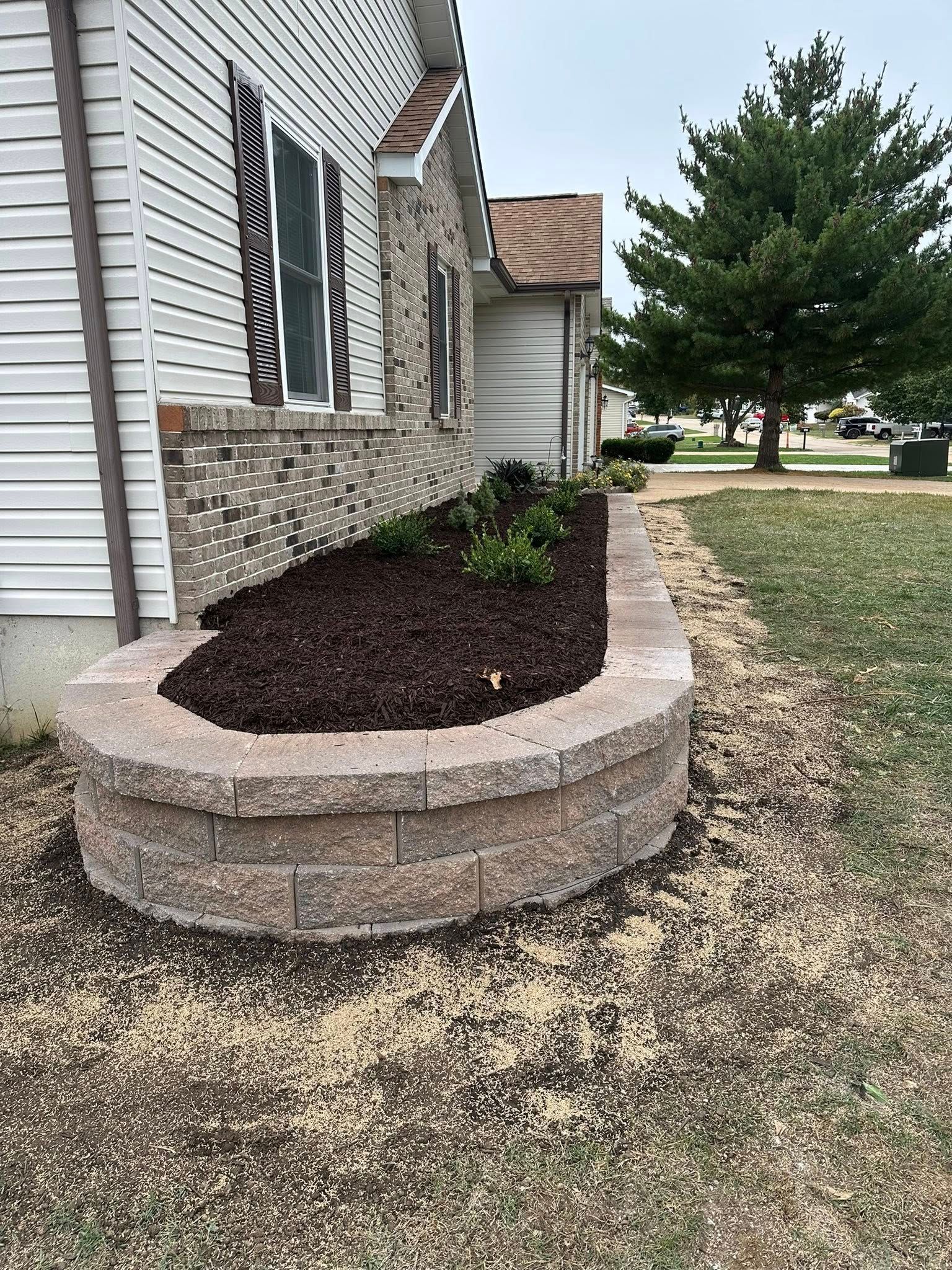 A raised garden bed with stone walls and fresh dark mulch, situated against the side of a house with light-colored siding. A raised garden bed with stone walls and fresh dark mulch, situated against the side of a house with light-colored siding.