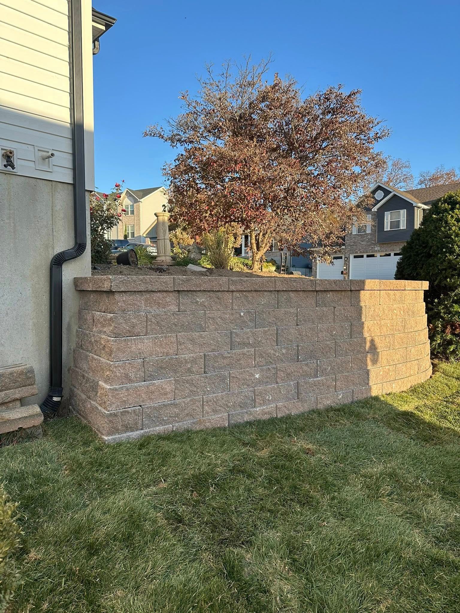 A tan stone retaining wall stands against the side of a house, set in a yard with a tree and blue sky background.