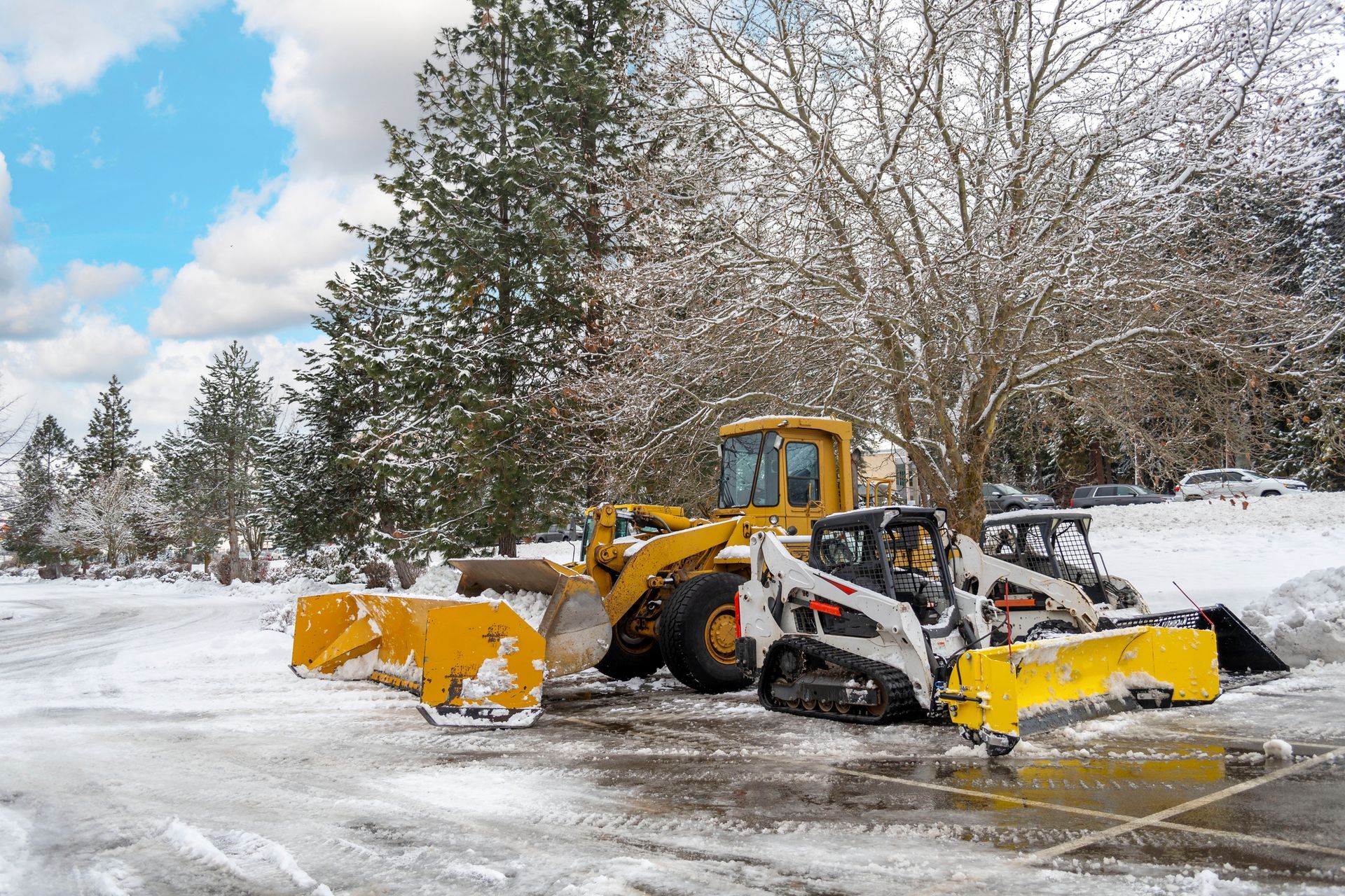 A yellow wheel loader and a white tracked skid steer with yellow snow blades sit on a snow-covered parking lot.
