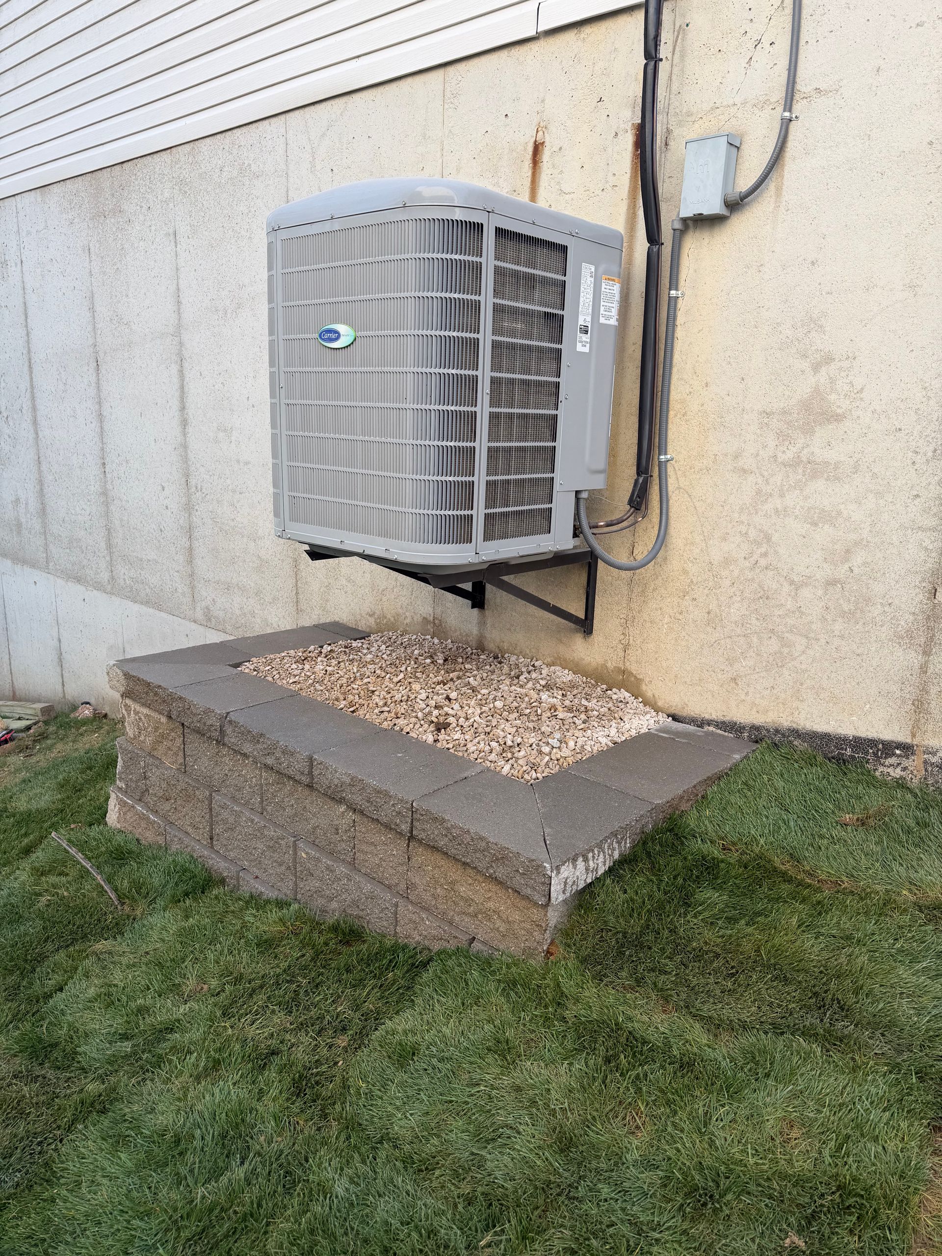 A light gray HVAC unit wall-mounted above a stone retaining wall filled with gravel next to a house exterior.