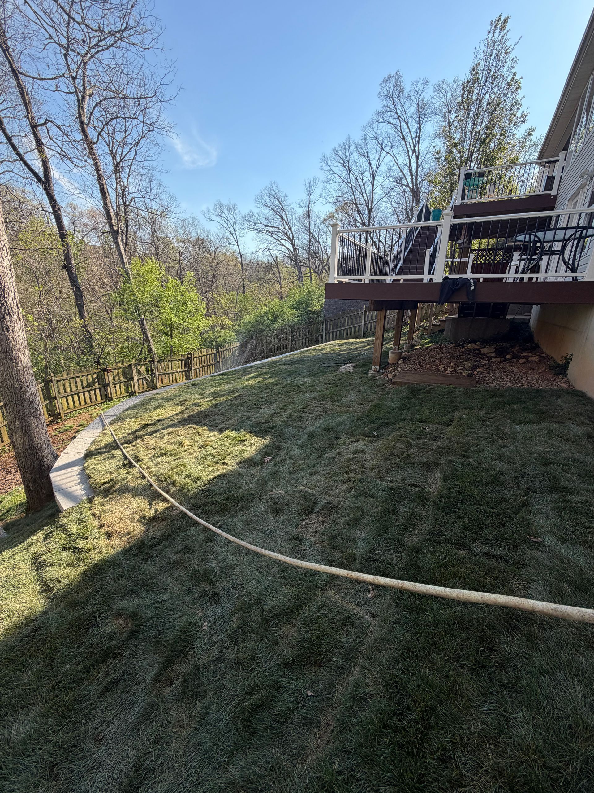 A sloping grassy lawn leads up to a wooden deck with a white railing under a clear blue sky, next to a wooded hillside.