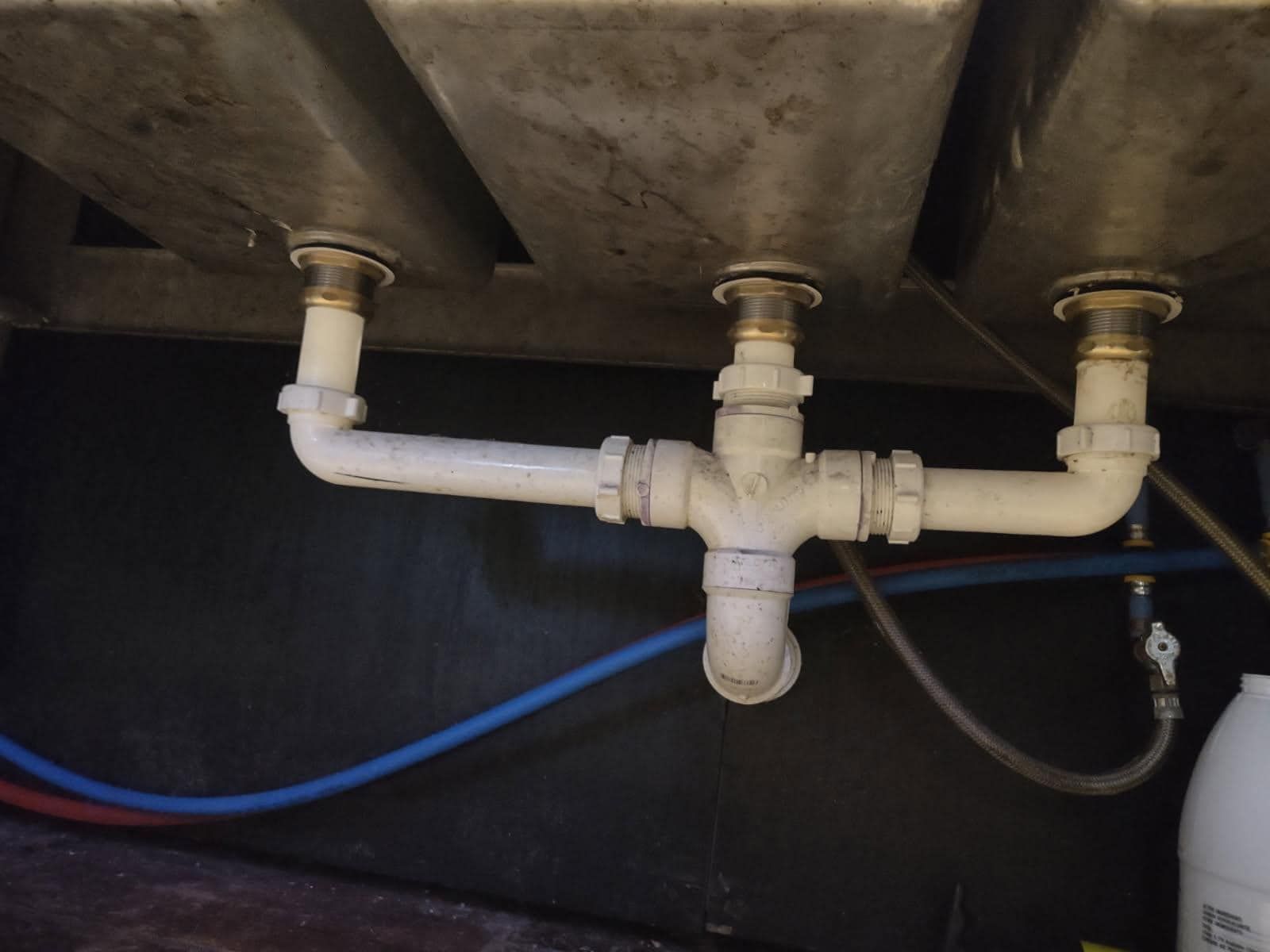 Underside of a three-basin sink. White PVC pipes connect the drains horizontally to a vertical drain pipe.
