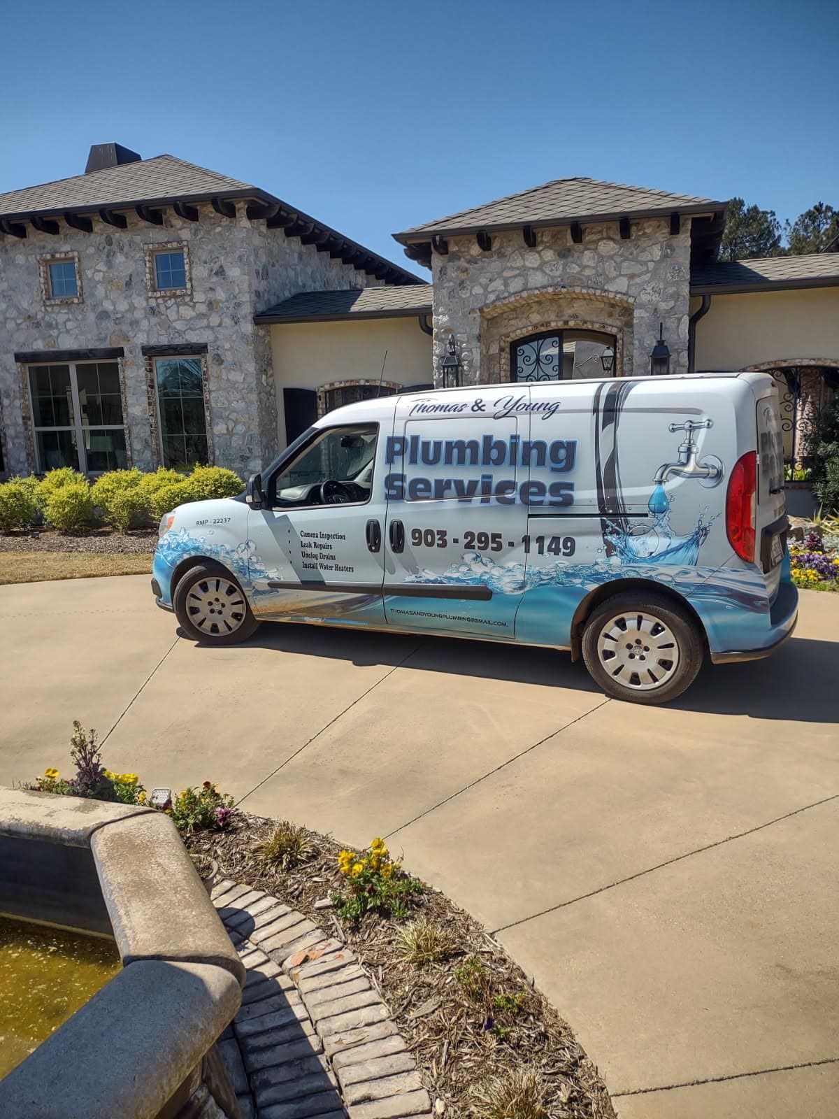 A plumbing services van parked in a driveway in front of a stone house on a sunny day.