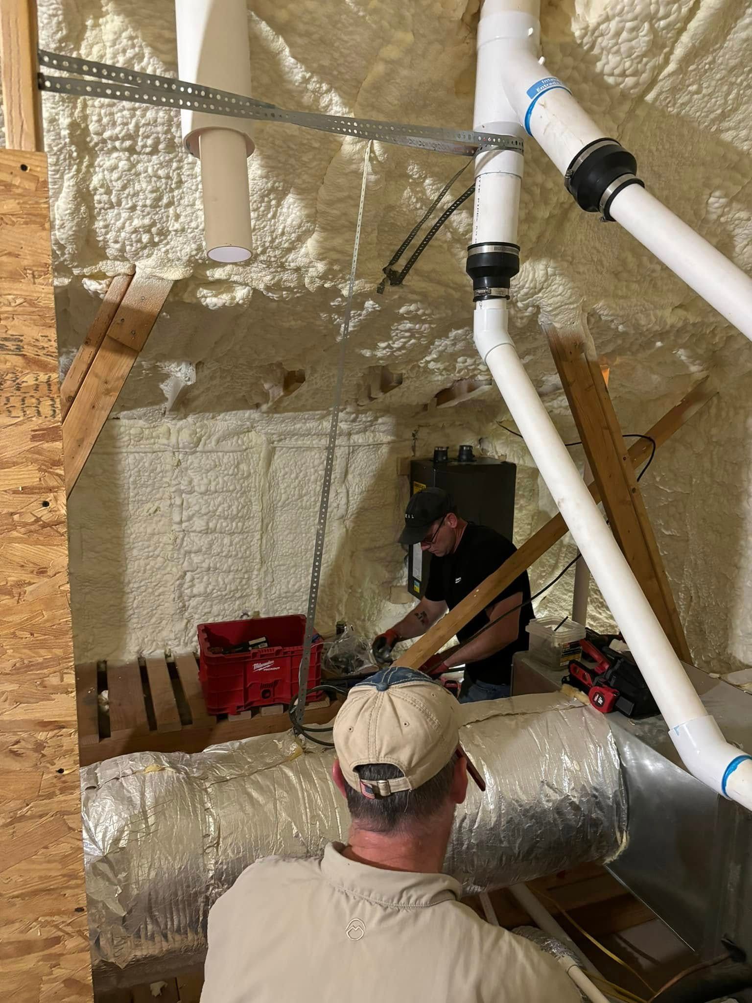 Two people working inside an attic with insulation and ductwork. One is cutting wood, the other is holding something.