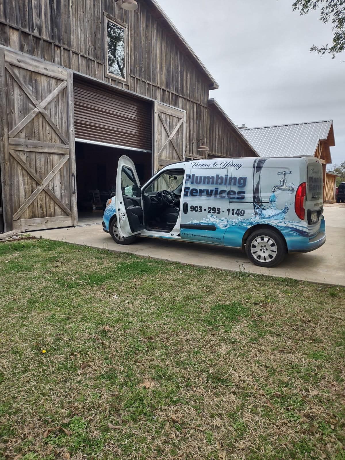 Plumbing service van parked in front of a barn. Van has open door and business logo. Green grass and cloudy sky.