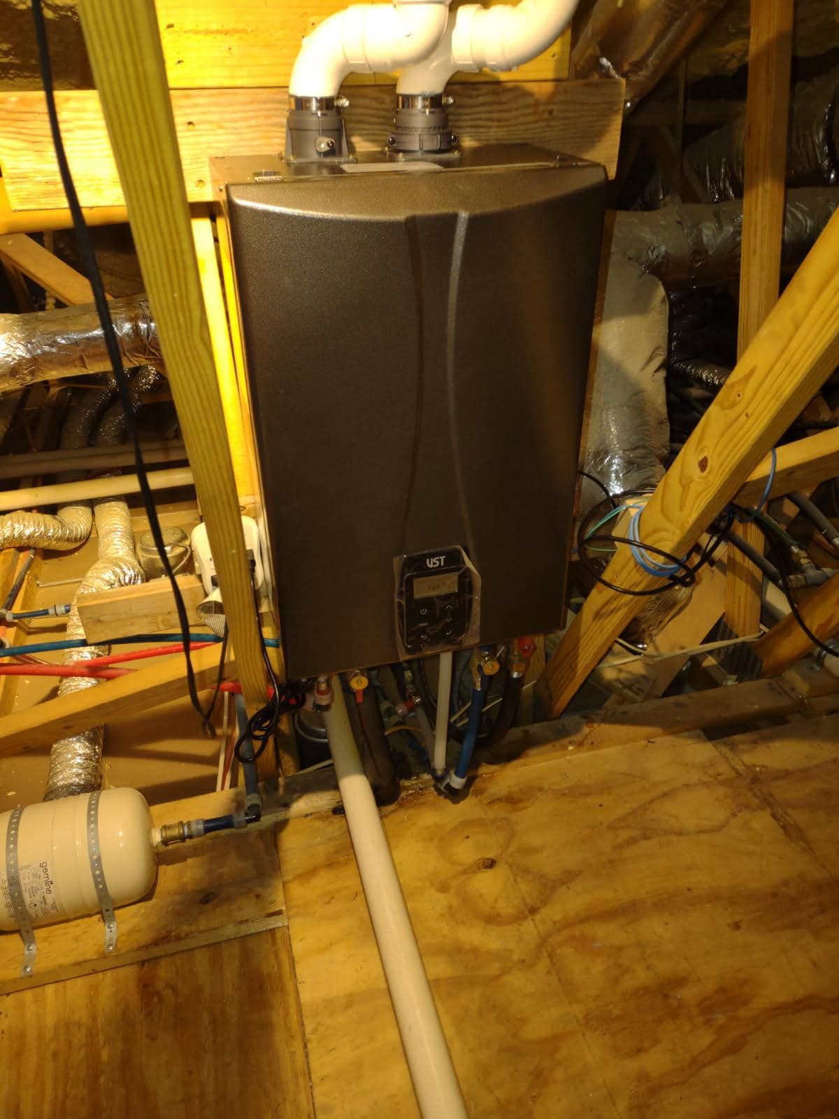A tankless water heater in an attic, with white pipes and wooden beams.
