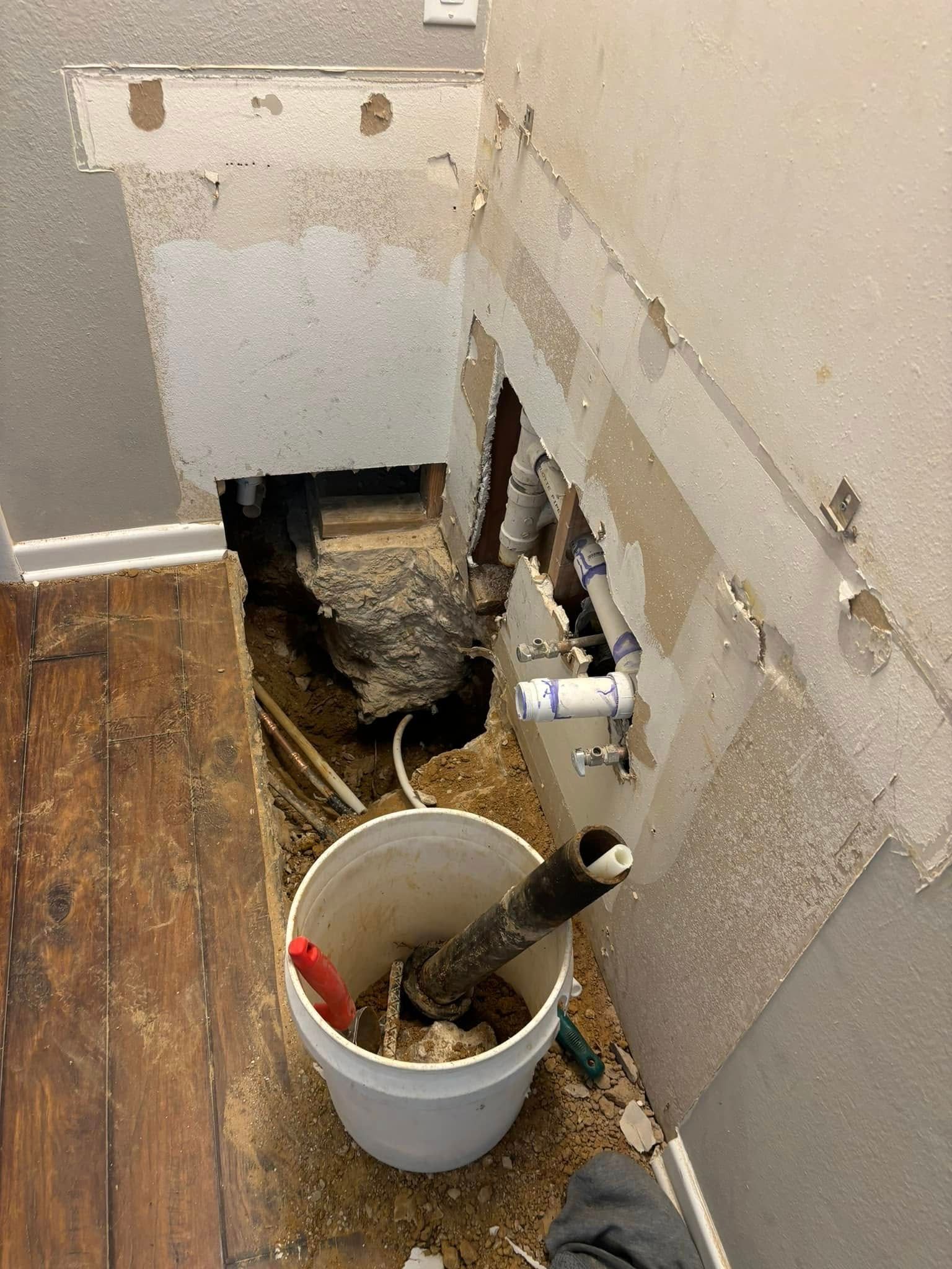 Bathroom wall undergoing repair; exposed pipes, bucket, debris, and torn drywall.