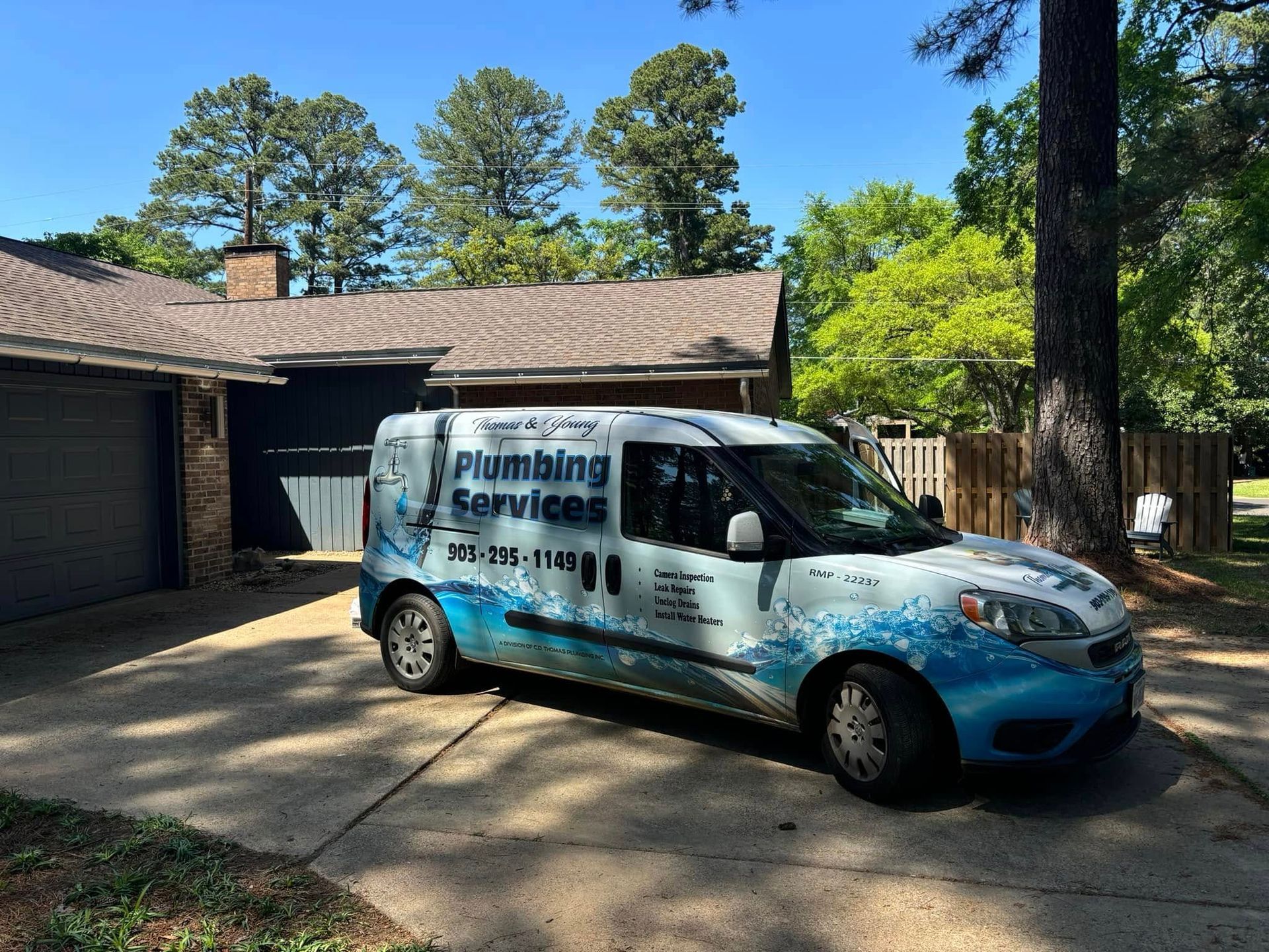 Plumbing services van parked in a driveway. Blue and white graphics, next to a garage and house, under a sunny sky.